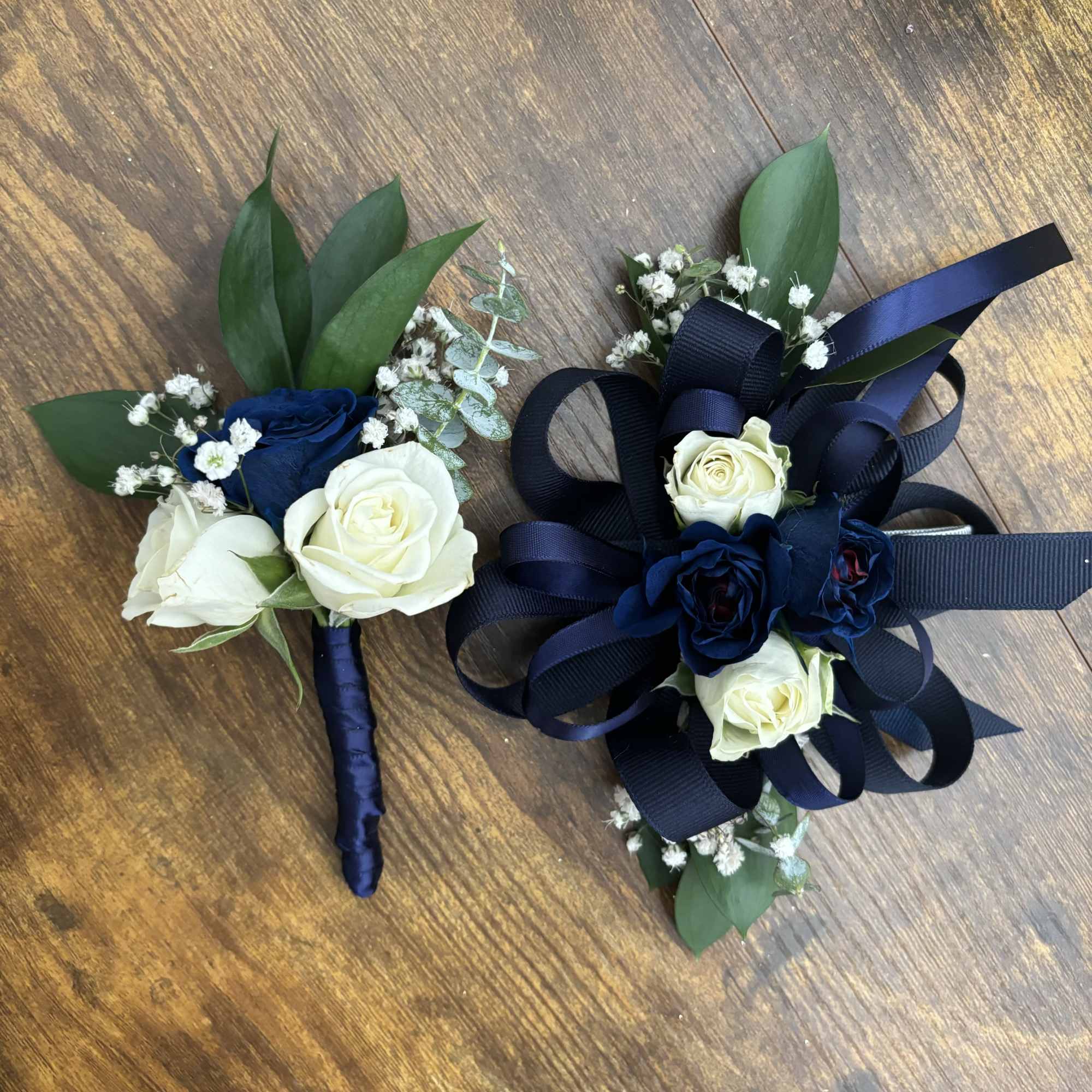 Two rose boutonnieres with navy ribbon and greenery on a wooden surface
