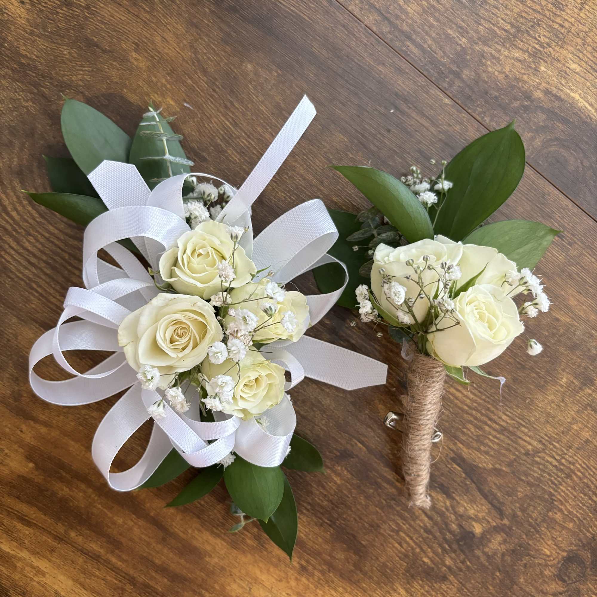 Two white rose boutonnieres with ribbon loops on a wooden surface