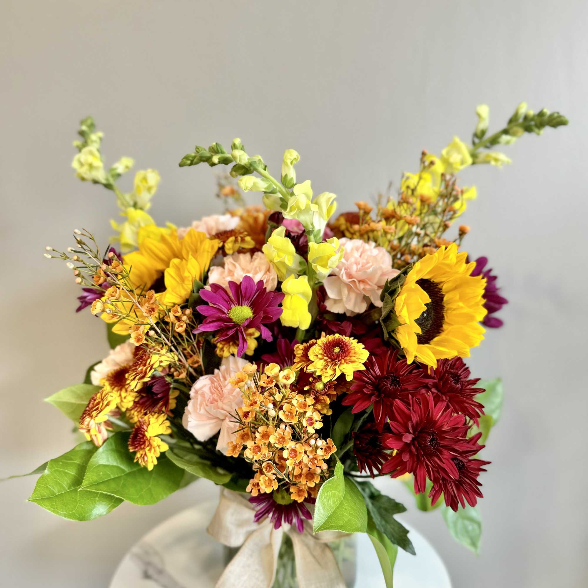 Bright mixed arrangement with sunflowers, burgundy mums, pink carnations, and yellow snapdragons in a glass vase