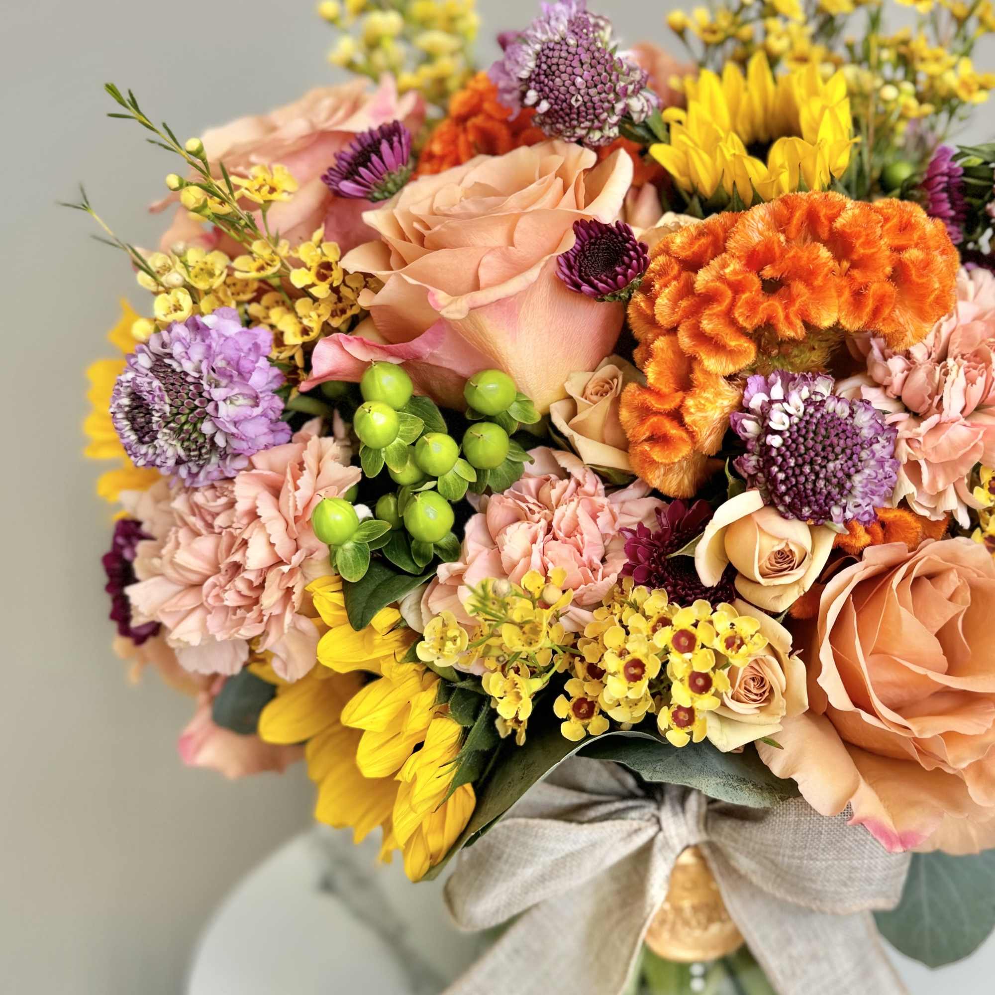 Vibrant mixed bouquet of yellow sunflowers, peach roses, and pink blooms in a clear vase with a fabric bow.