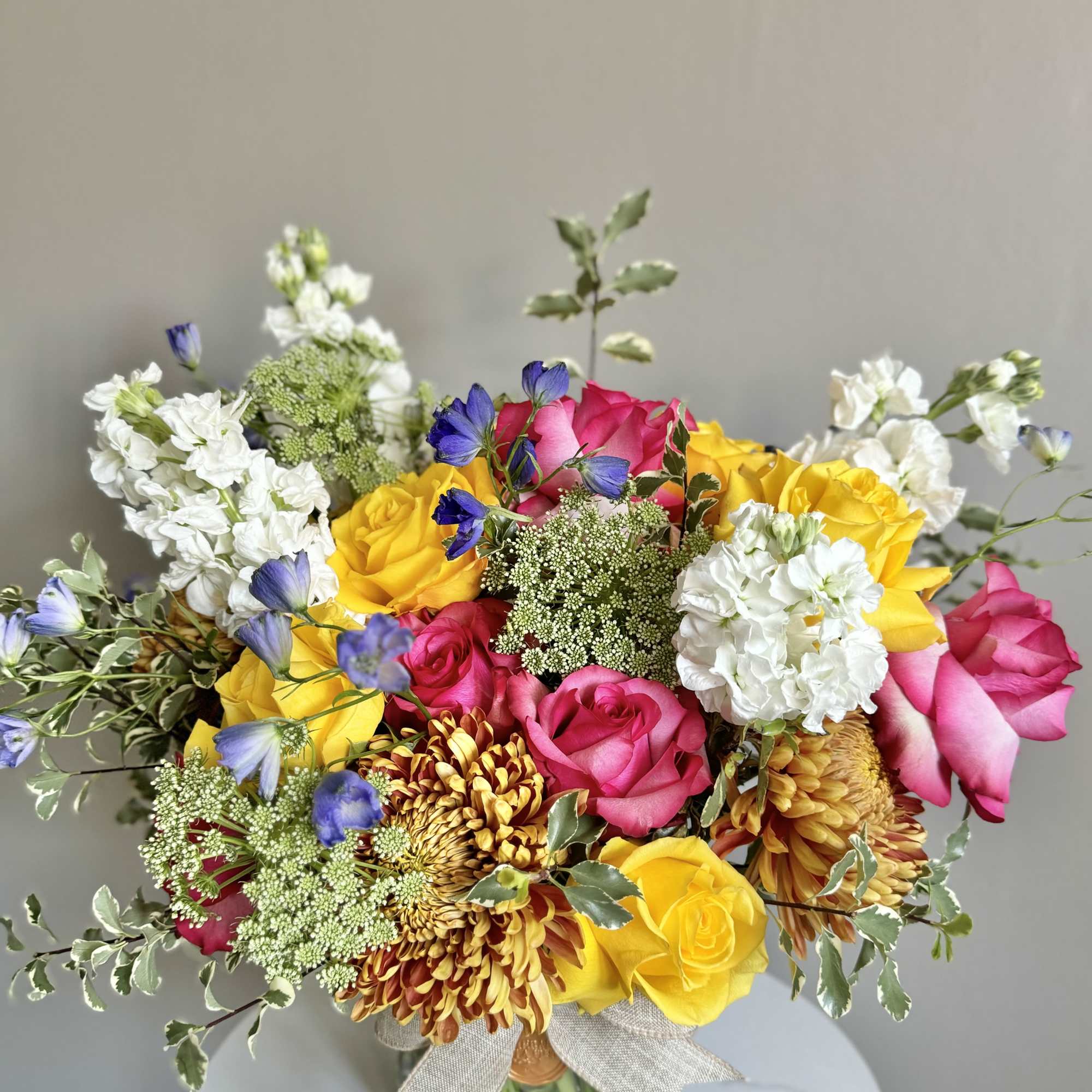Bright arrangement of yellow and pink roses, white stock, bronze mums, and blue accents in a glass vase with a bow