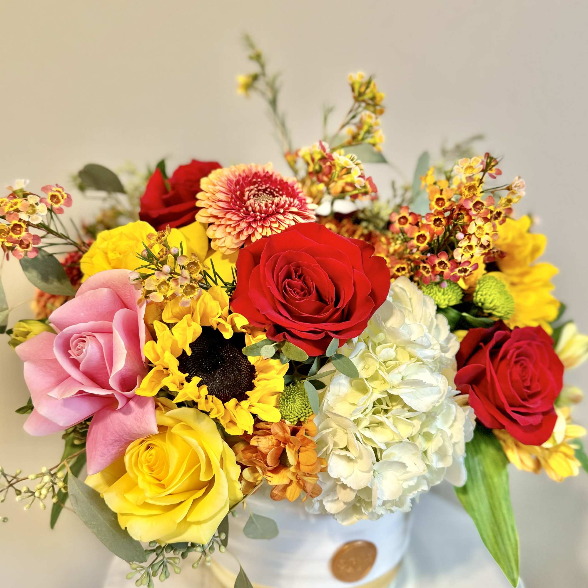 Bright arrangement of red, yellow, and pink roses with sunflowers and hydrangea in a white hatbox container