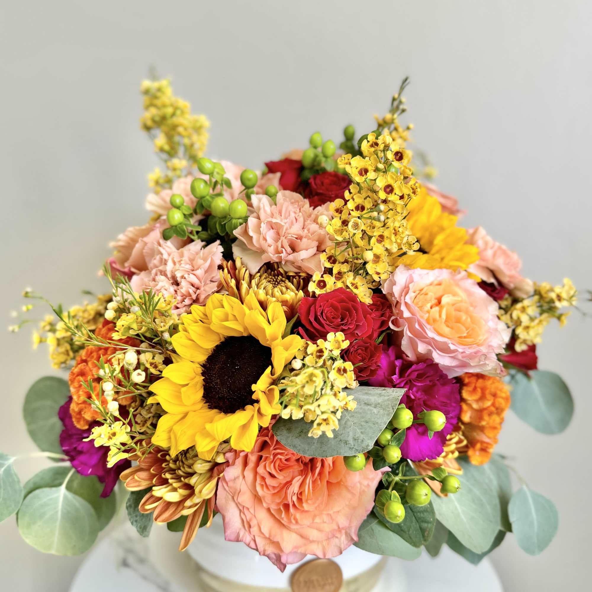 Bright arrangement with sunflowers, roses, carnations, and orchids in a white round container
