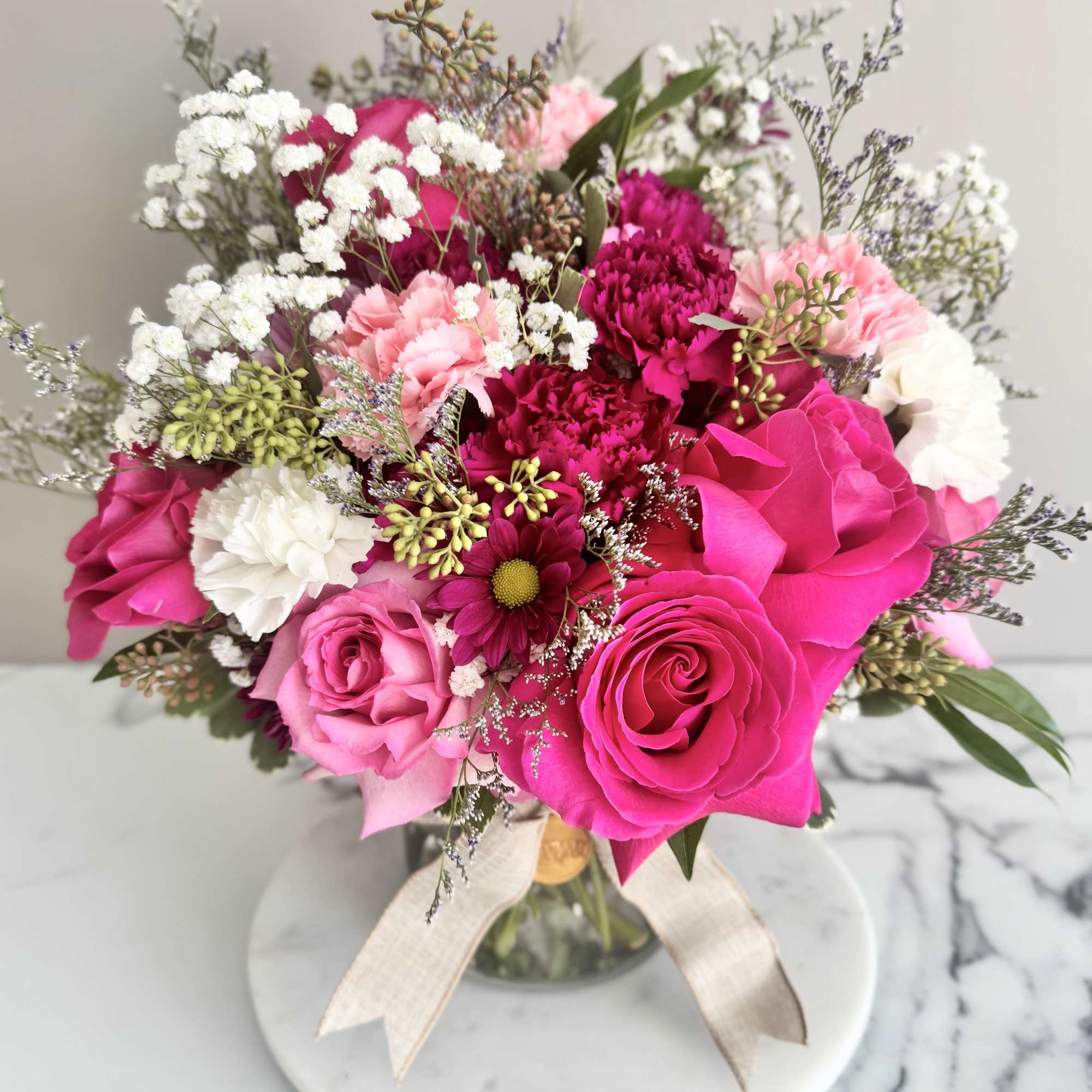 Compact bouquet of hot pink roses, pink carnations, and white blooms in a glass vase with a ribbon.