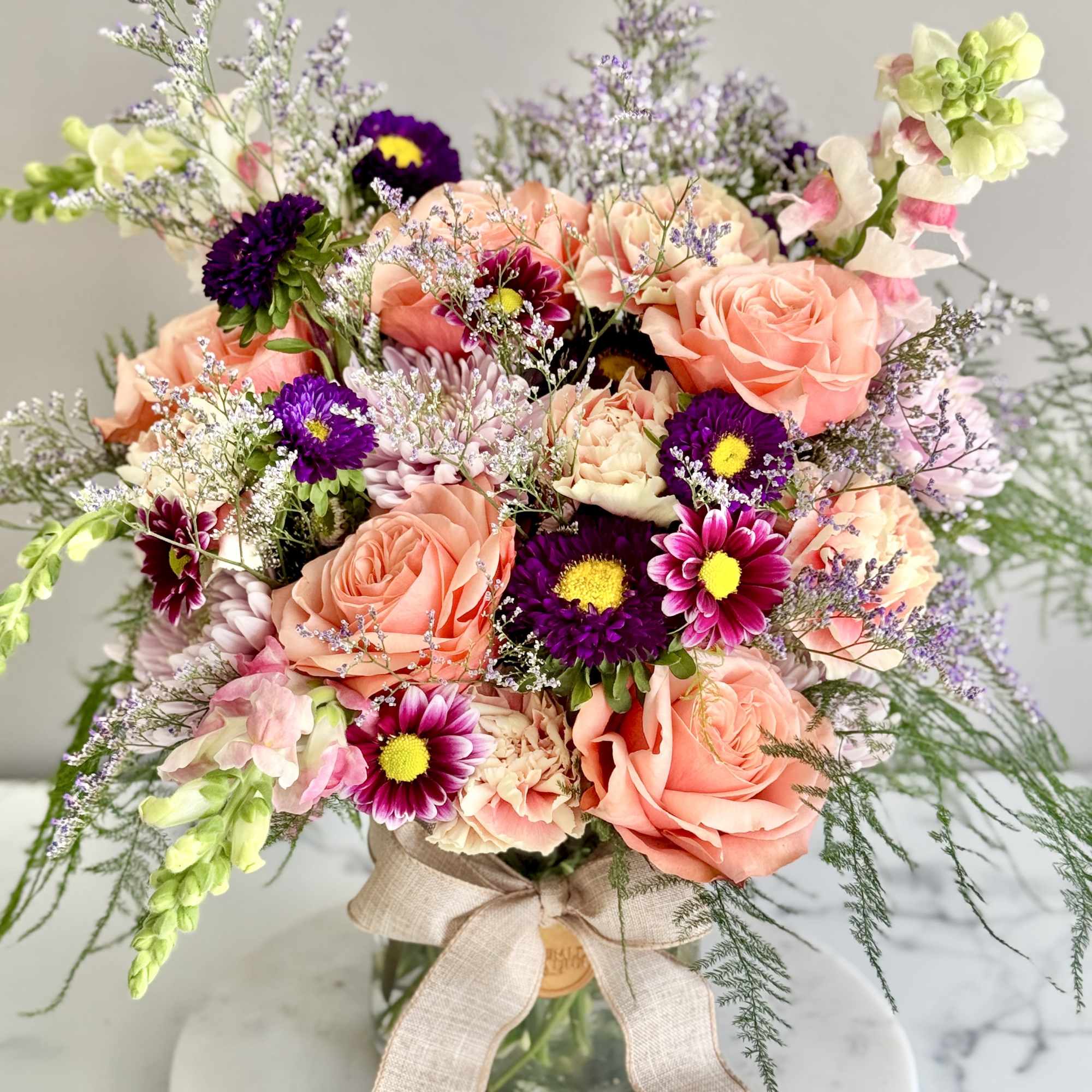 Peach roses and purple asters with mixed blooms in a glass jar vase tied with a linen bow