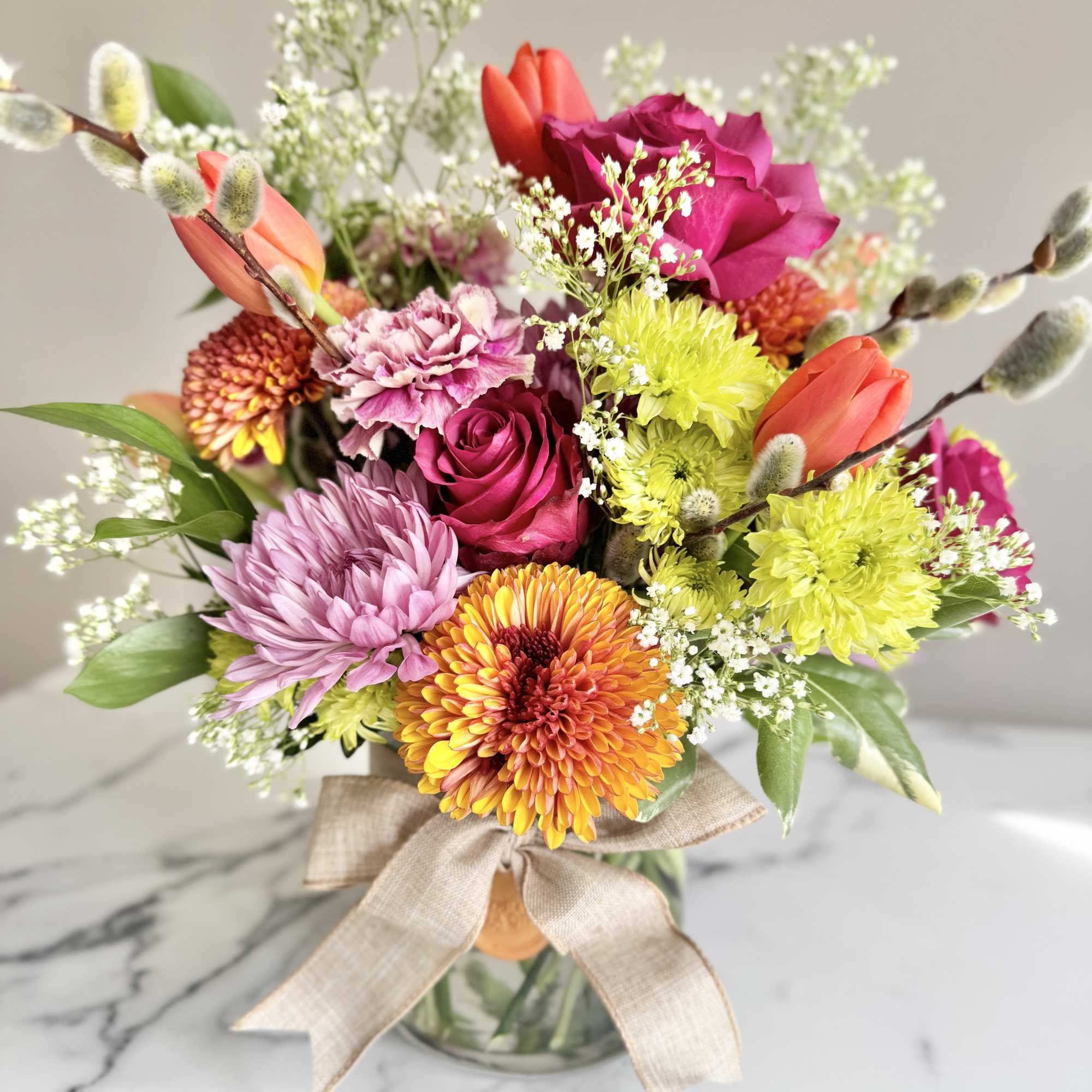 Bright mixed bouquet of roses, tulips, mums, and carnations in a glass vase with a bow