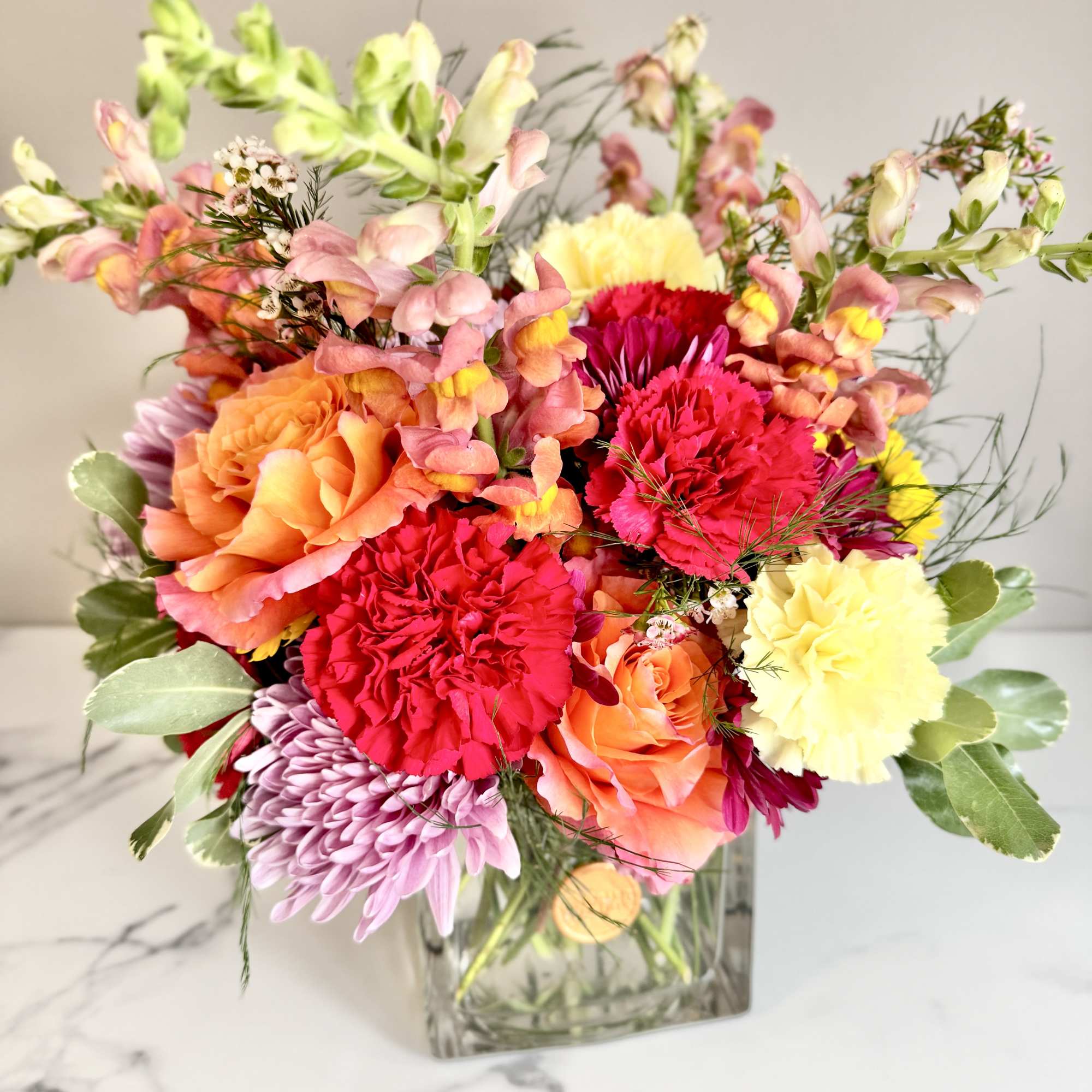 Bright mixed bouquet of roses, carnations, mums, and snapdragons in a clear glass cube vase