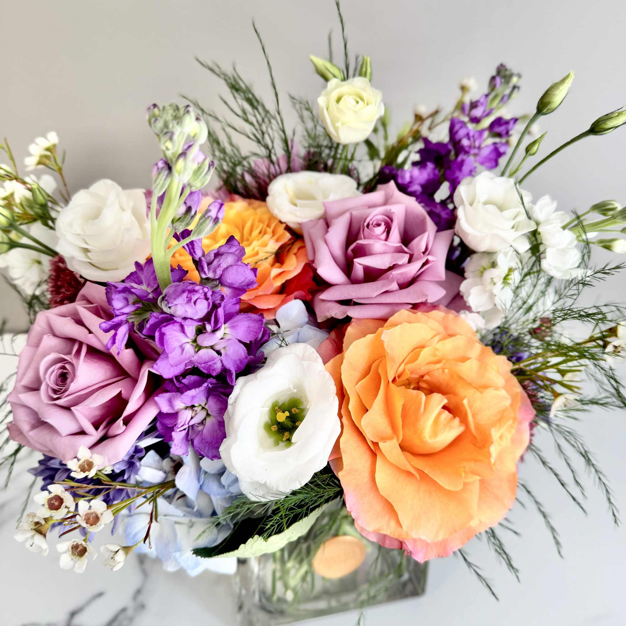 Colorful arrangement of orange and lavender roses with white lisianthus and purple blooms in a clear glass cube vase