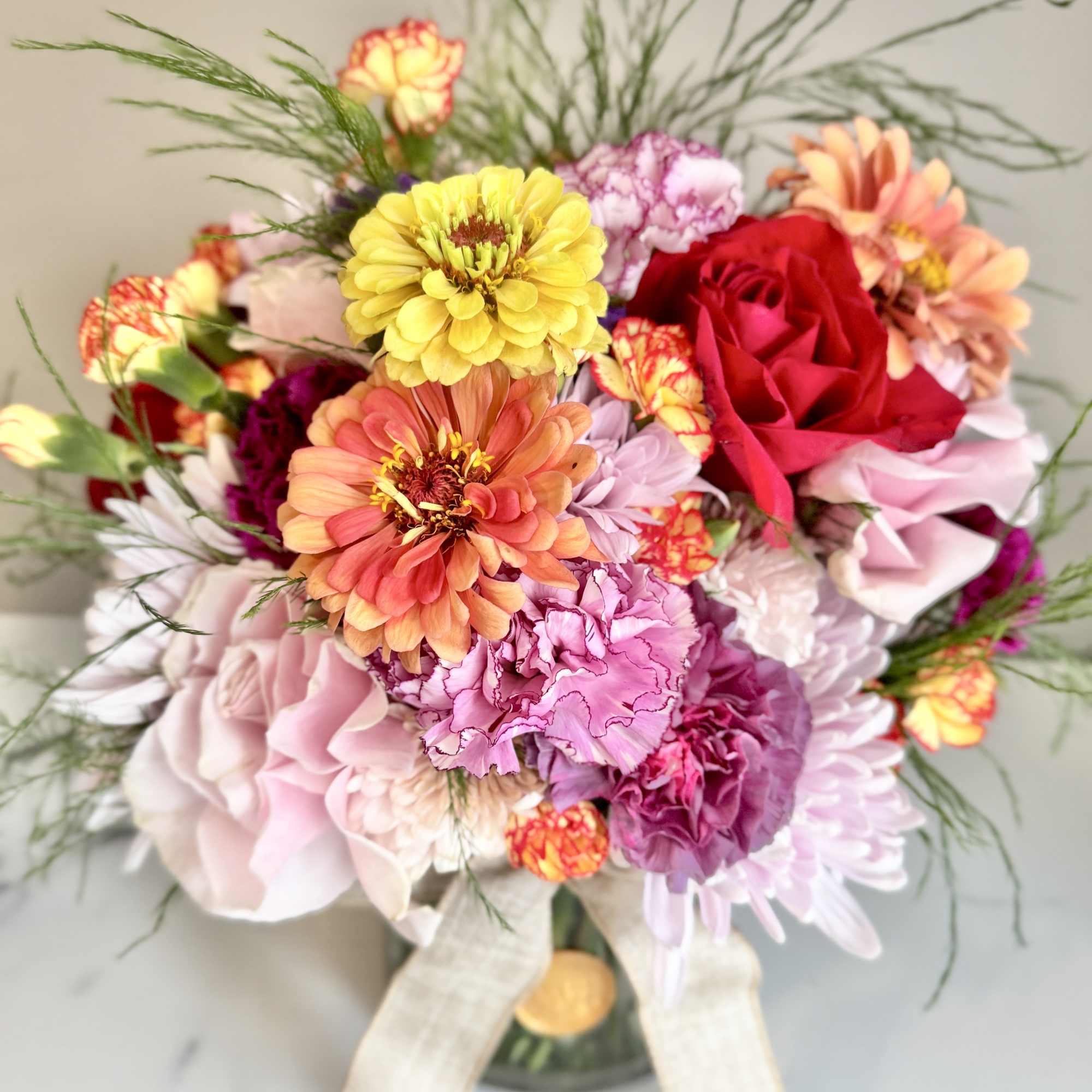 Bright mixed arrangement of pink, yellow, and orange blooms in a clear glass vase with a linen bow.