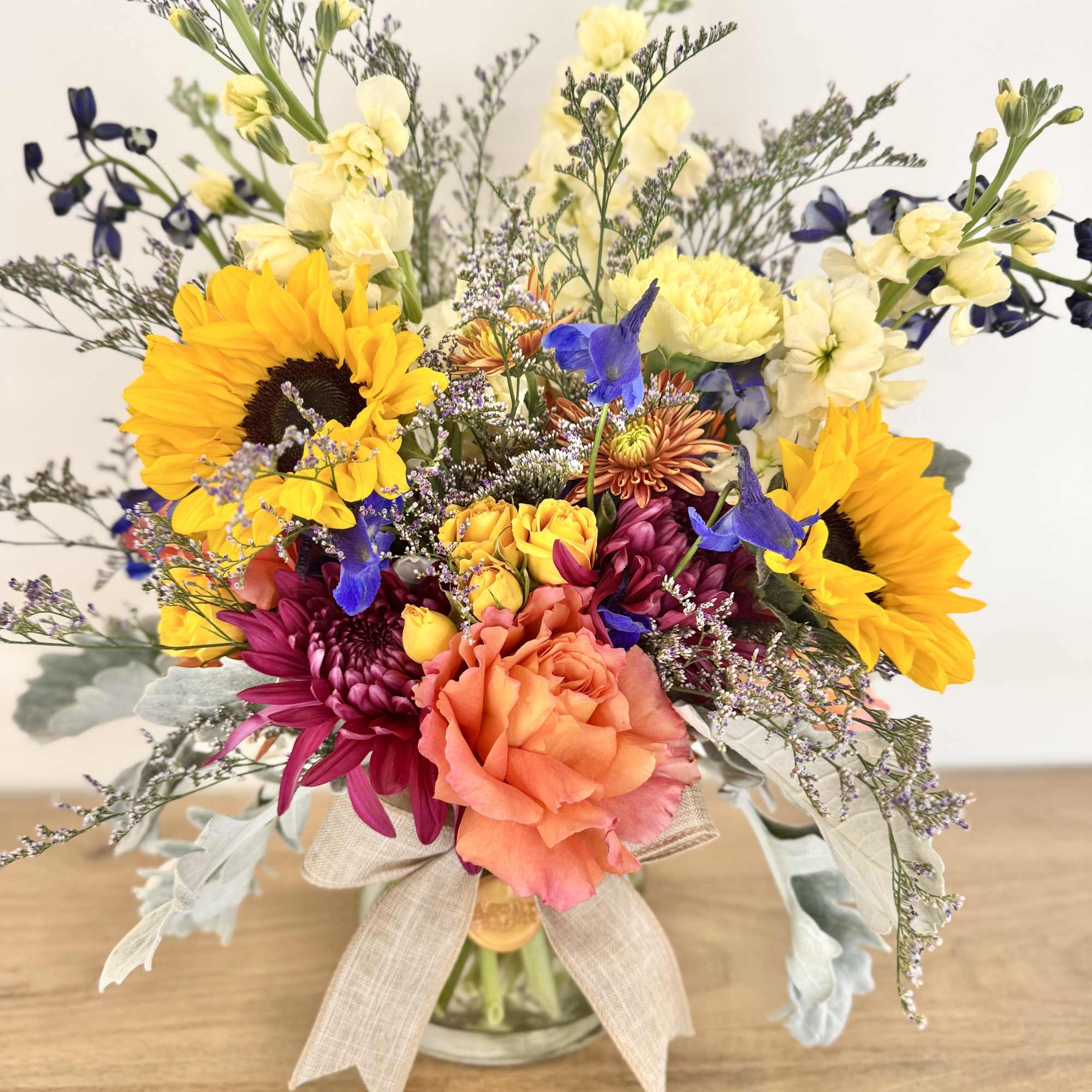 Bright mixed bouquet with sunflowers, orange rose, and purple blooms in a glass jar with a bow