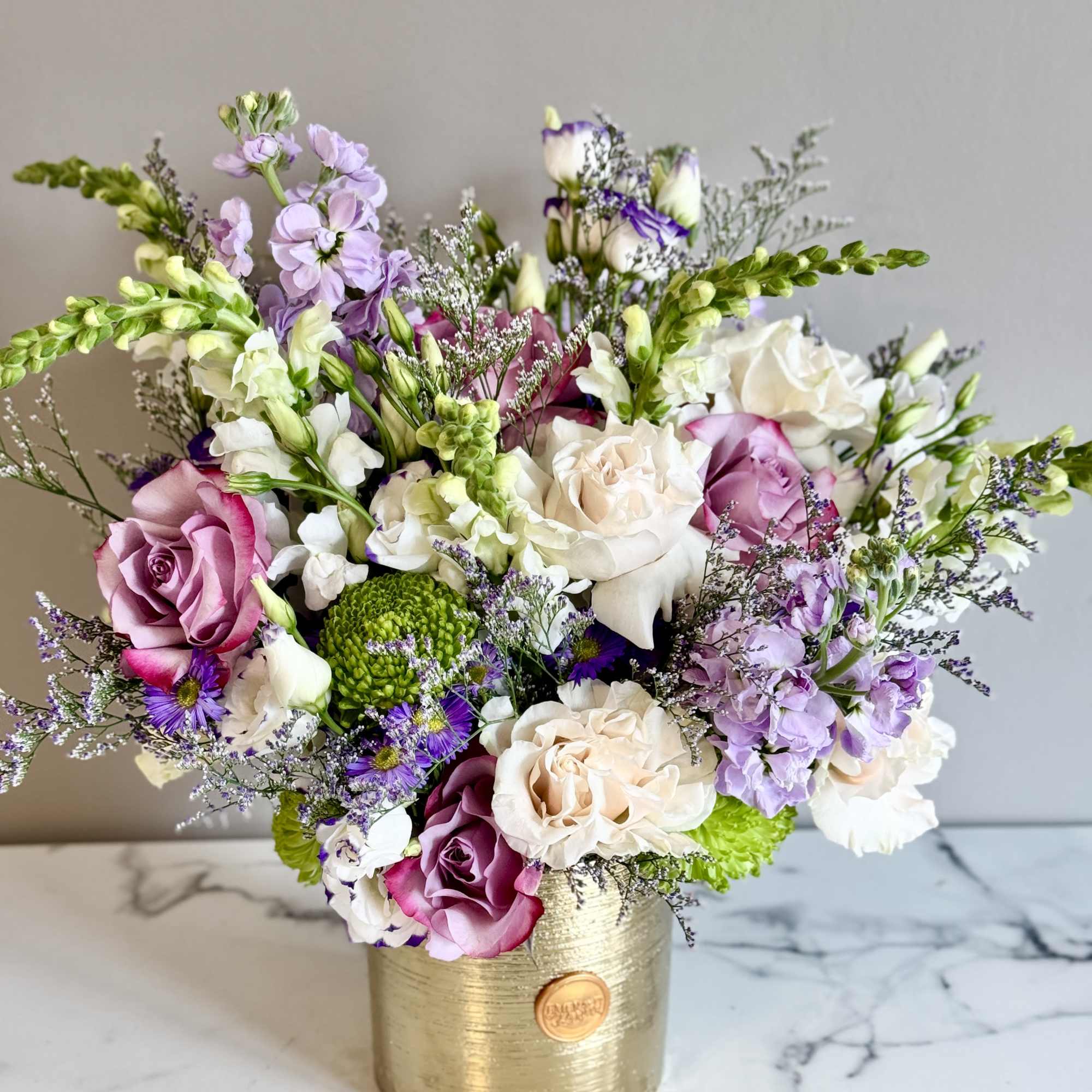 Lavender, white, and pink mixed blooms arranged in a gold cylinder vase on a marble surface.