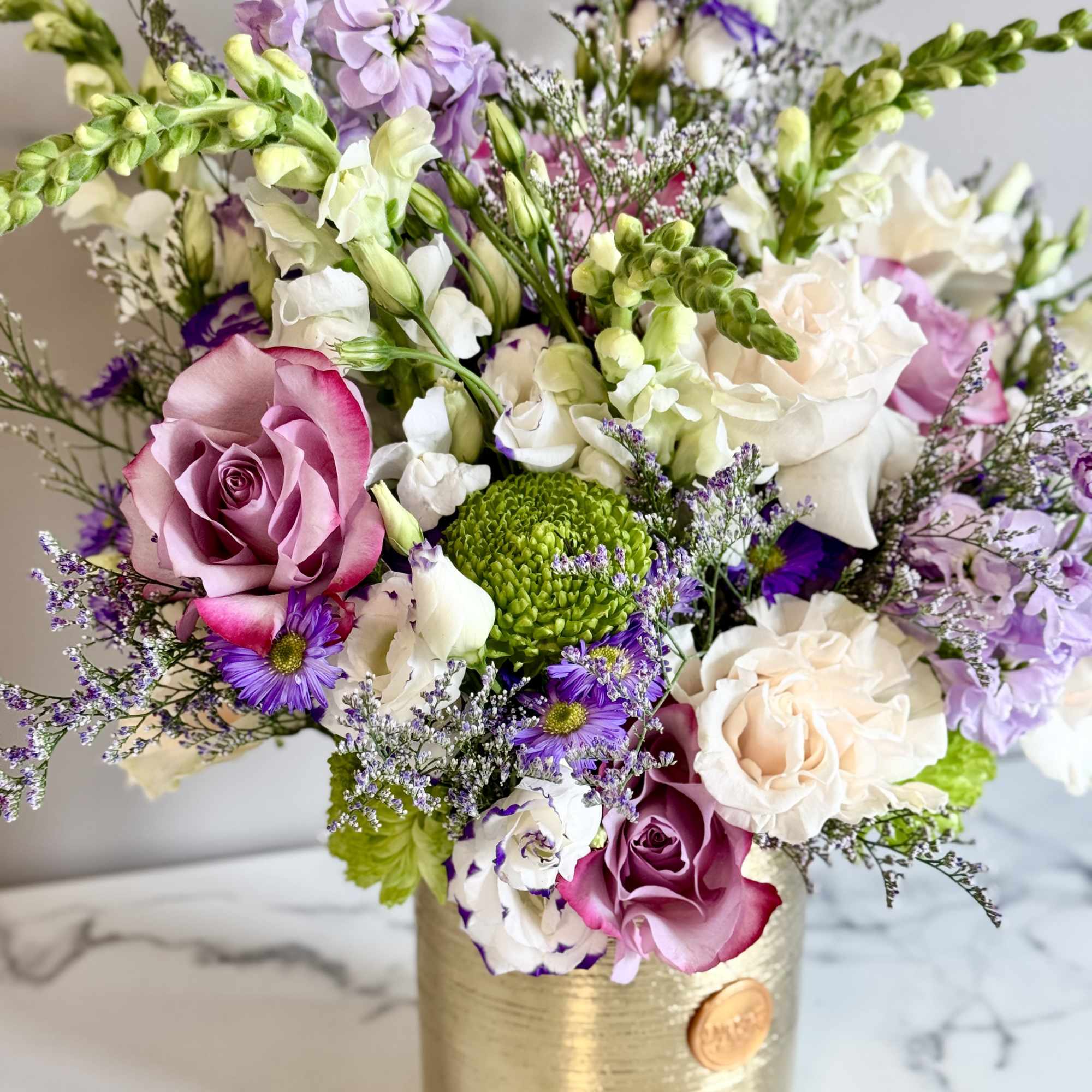 Mixed arrangement of pink roses, white blooms, and purple flowers in a gold cylinder vase