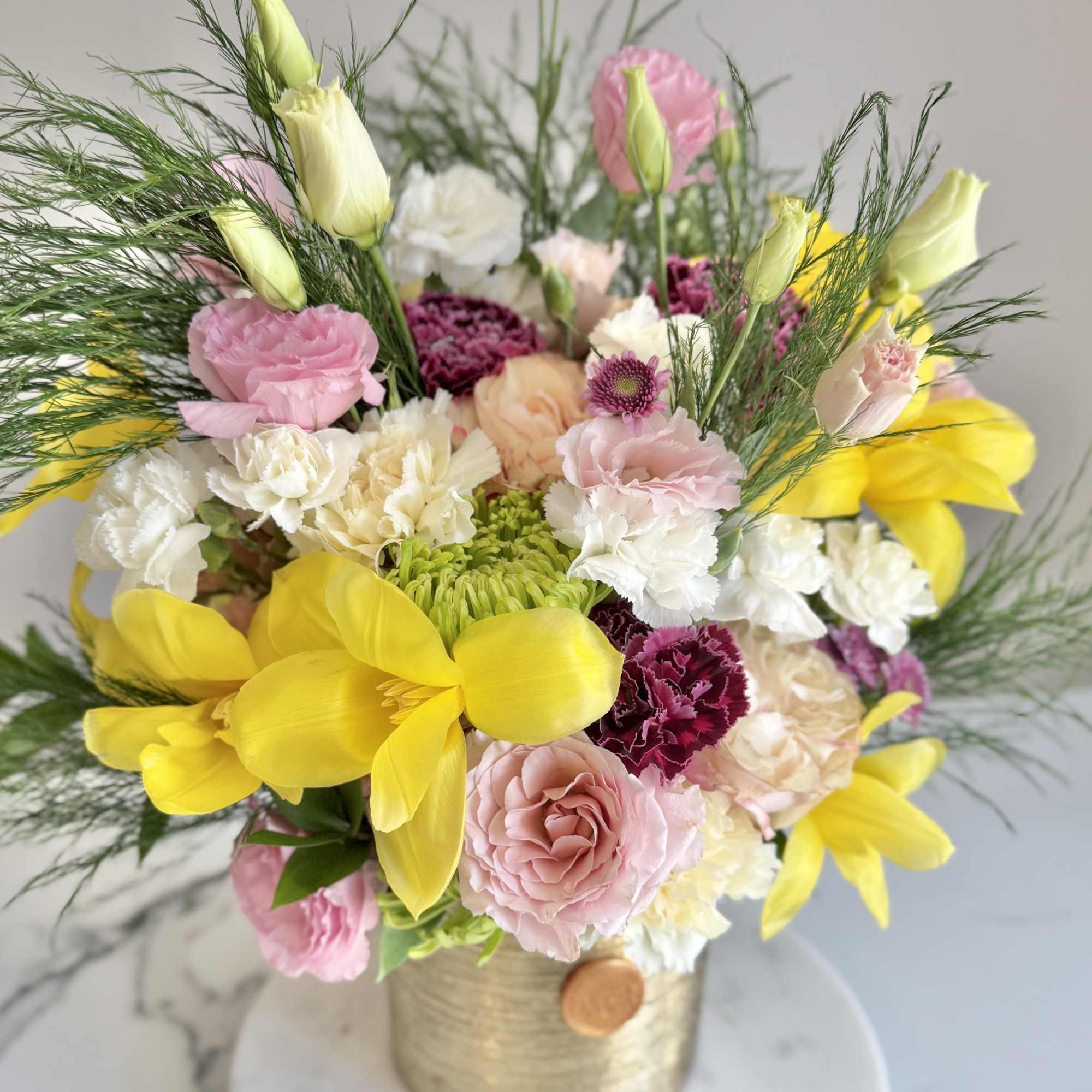 Colorful arrangement of yellow lilies, pink lisianthus, and mixed blooms in a gold cylinder vase