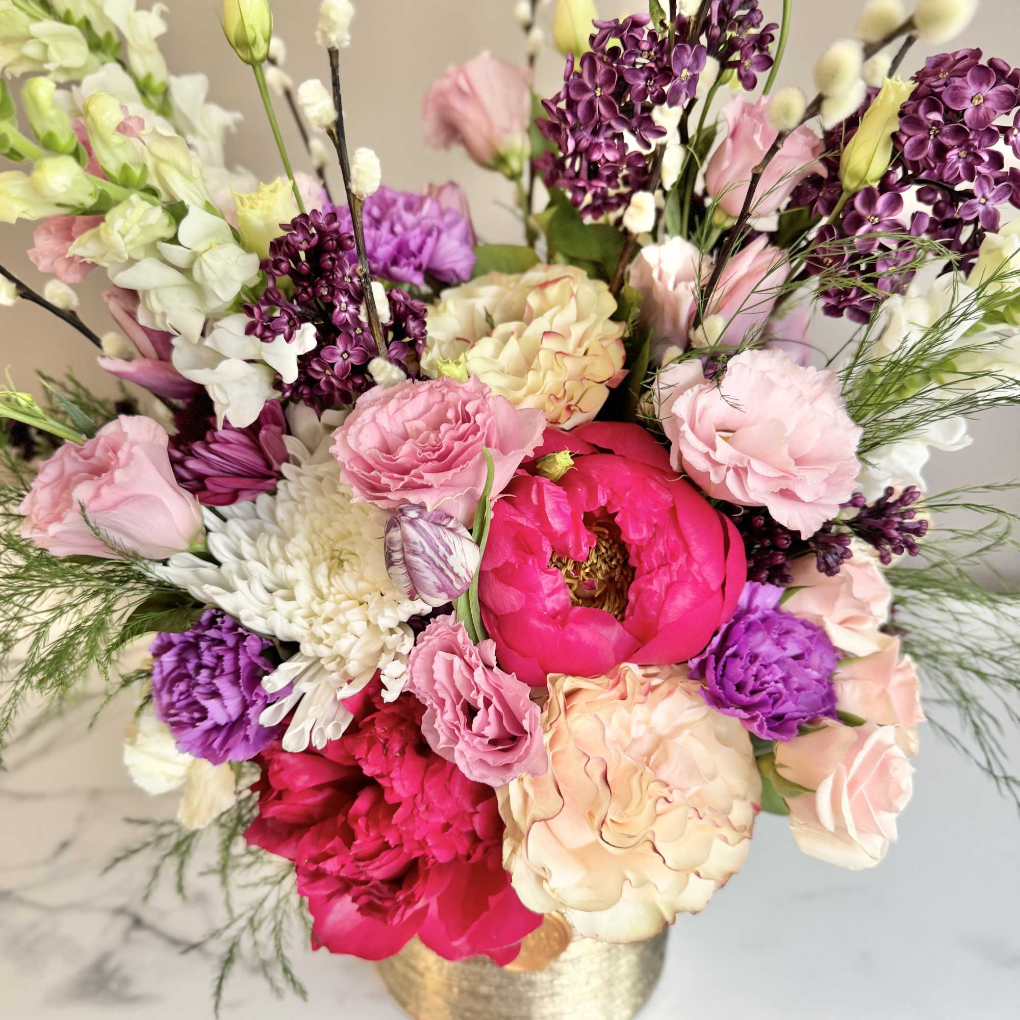 Mixed arrangement of pink peonies, roses, lilac, and white mums in a gold cylinder vase