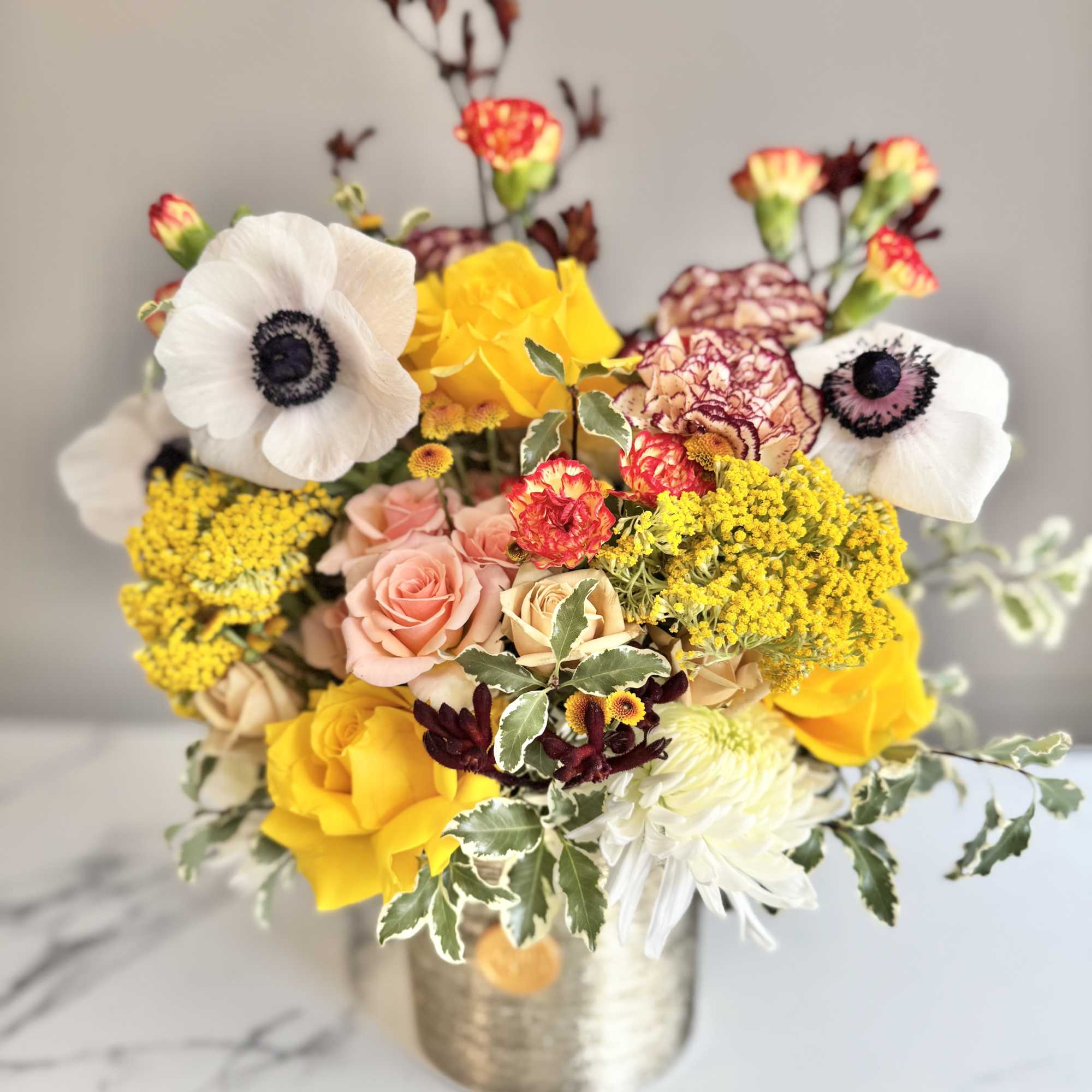 Colorful arrangement with yellow roses, white anemones, peach roses and carnations in a gold metal vase