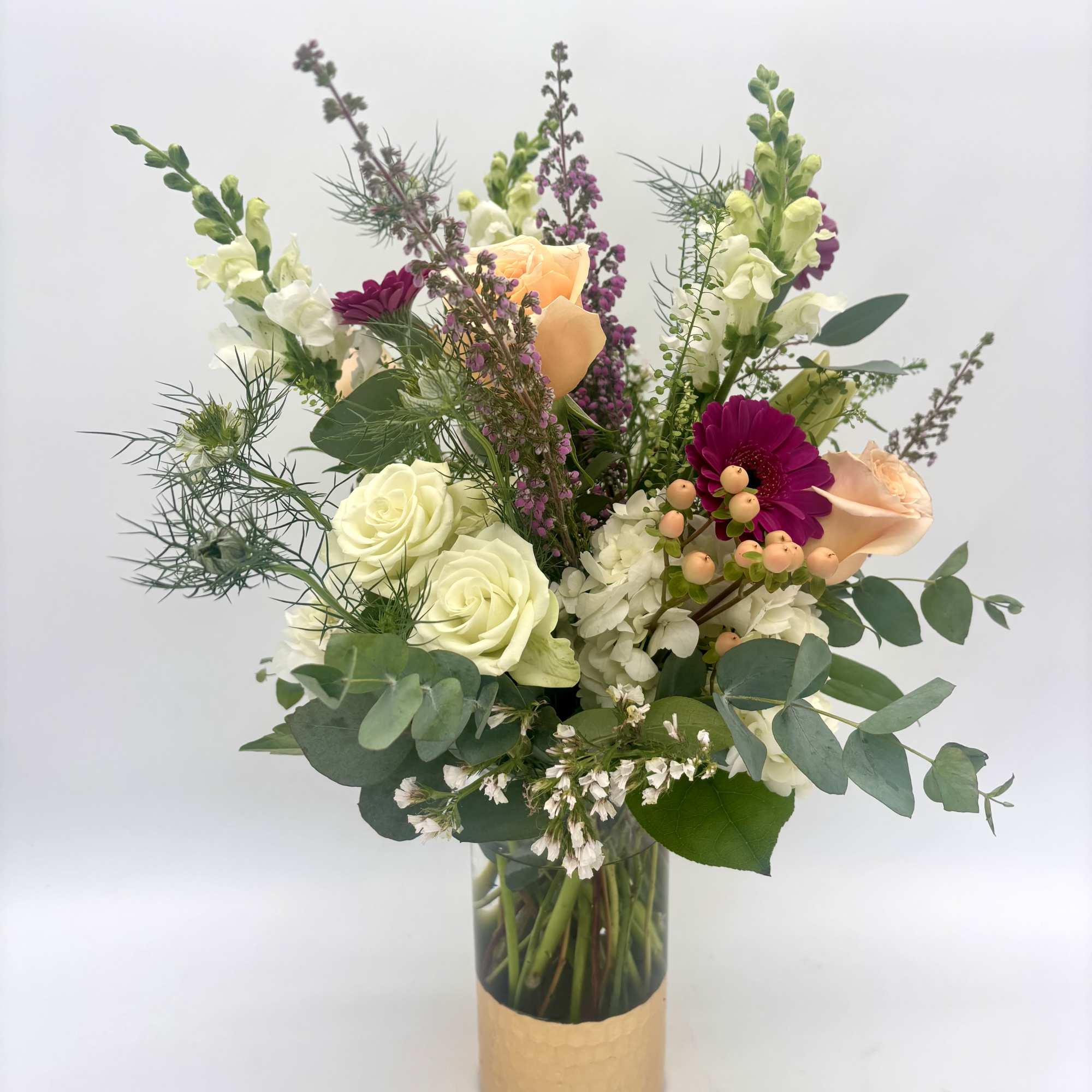 Tall mixed bouquet of white and peach roses, magenta daisies, and snapdragons in a glass vase with gold base