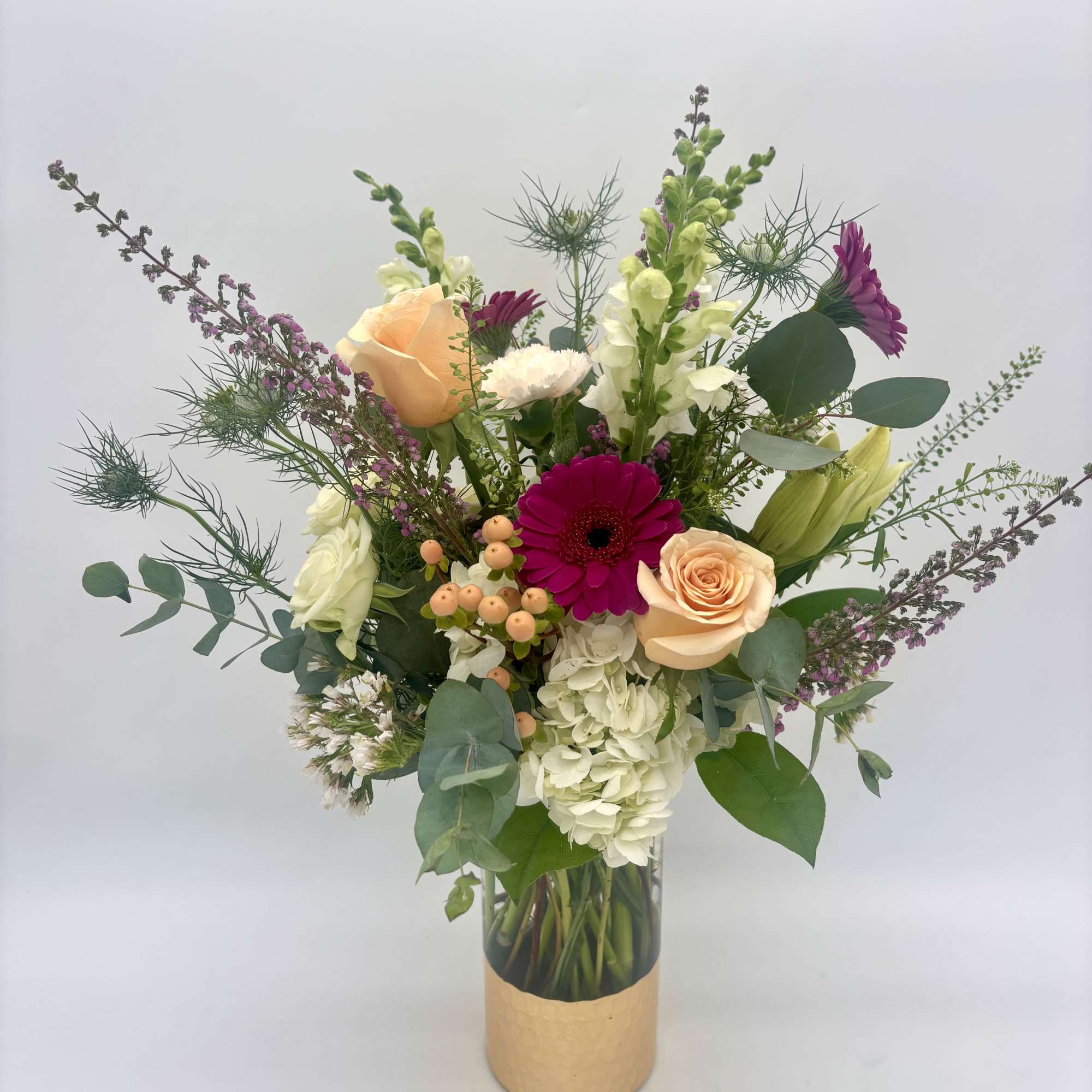 Tall mixed bouquet of white hydrangea, peach roses, and magenta gerbera daisies in a glass vase with a gold band.