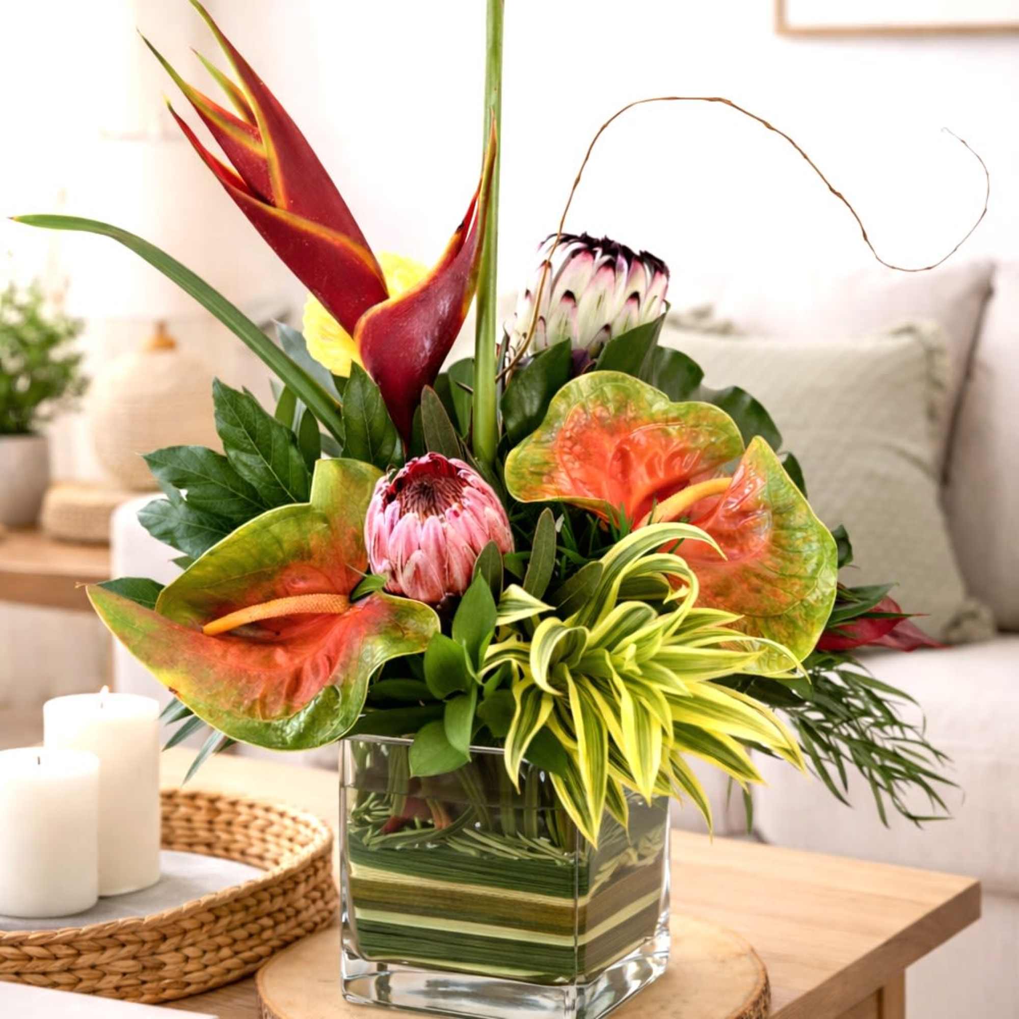 Tropical arrangement with heliconia, protea, and anthurium in a square glass vase on a coffee table.