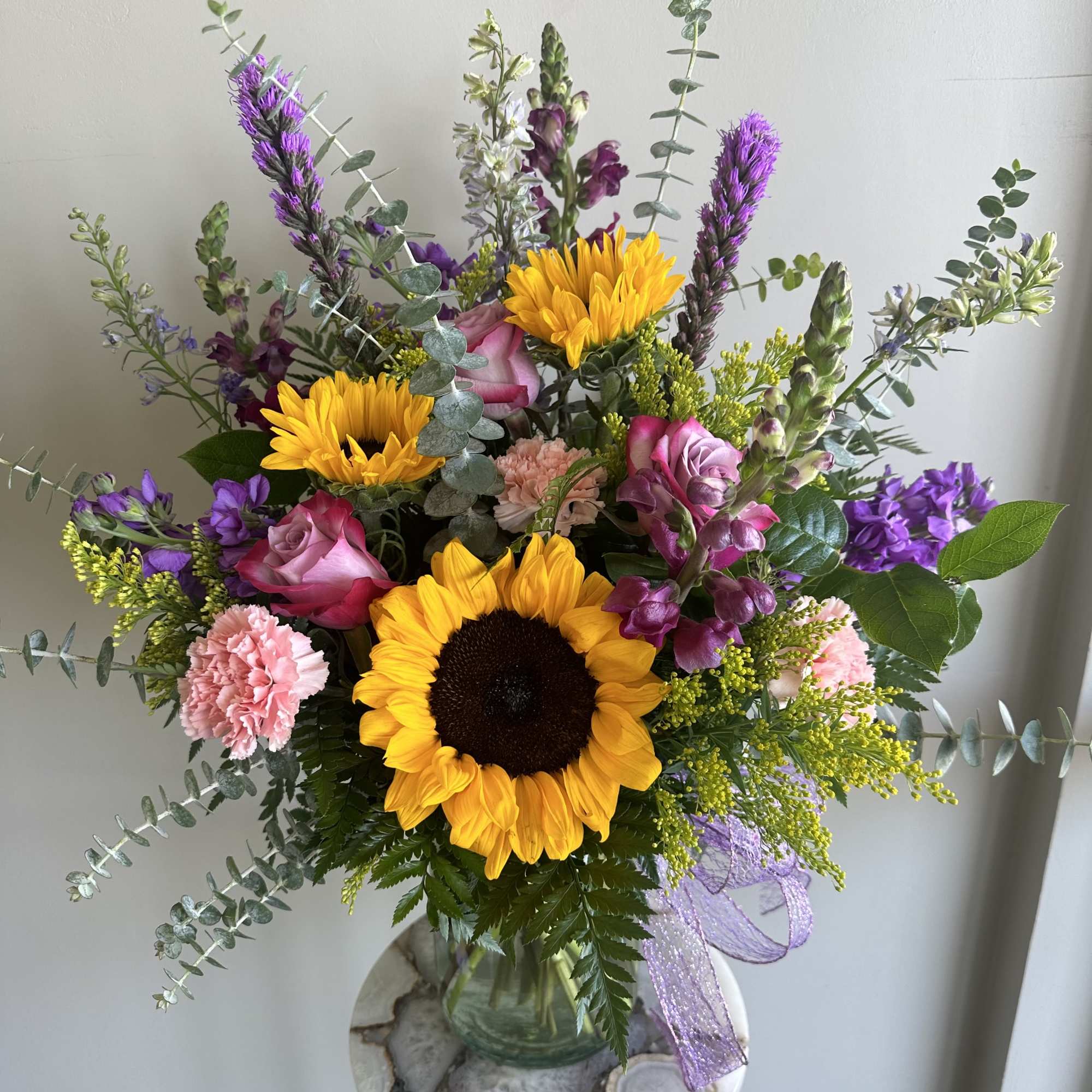 Mixed bouquet of sunflowers, pink roses, and purple flowers in a clear glass vase with ribbon