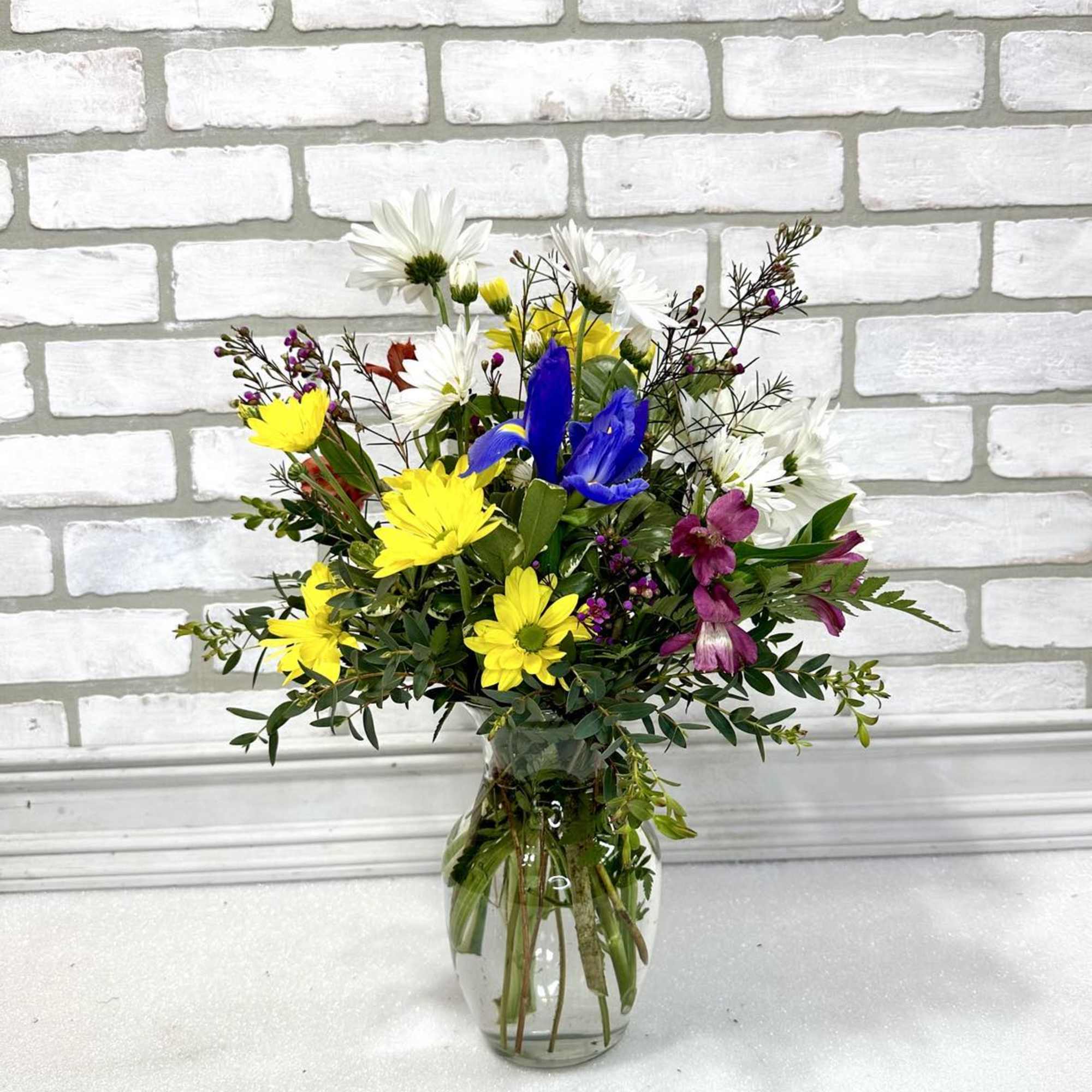 Mixed bouquet of yellow, white, blue, and purple flowers in a clear glass vase
