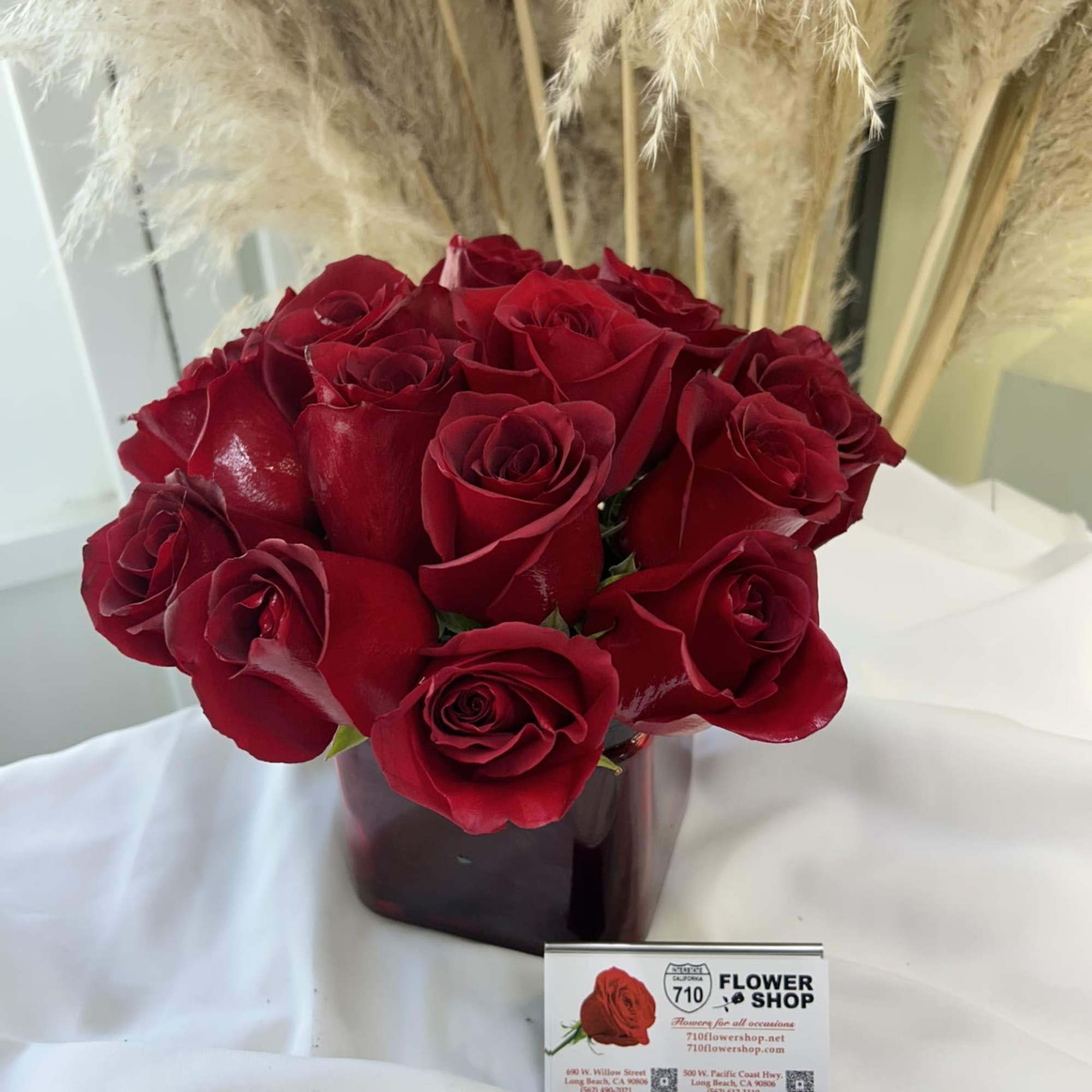 Red roses arranged in a dark vase with tall pampas grass behind them.