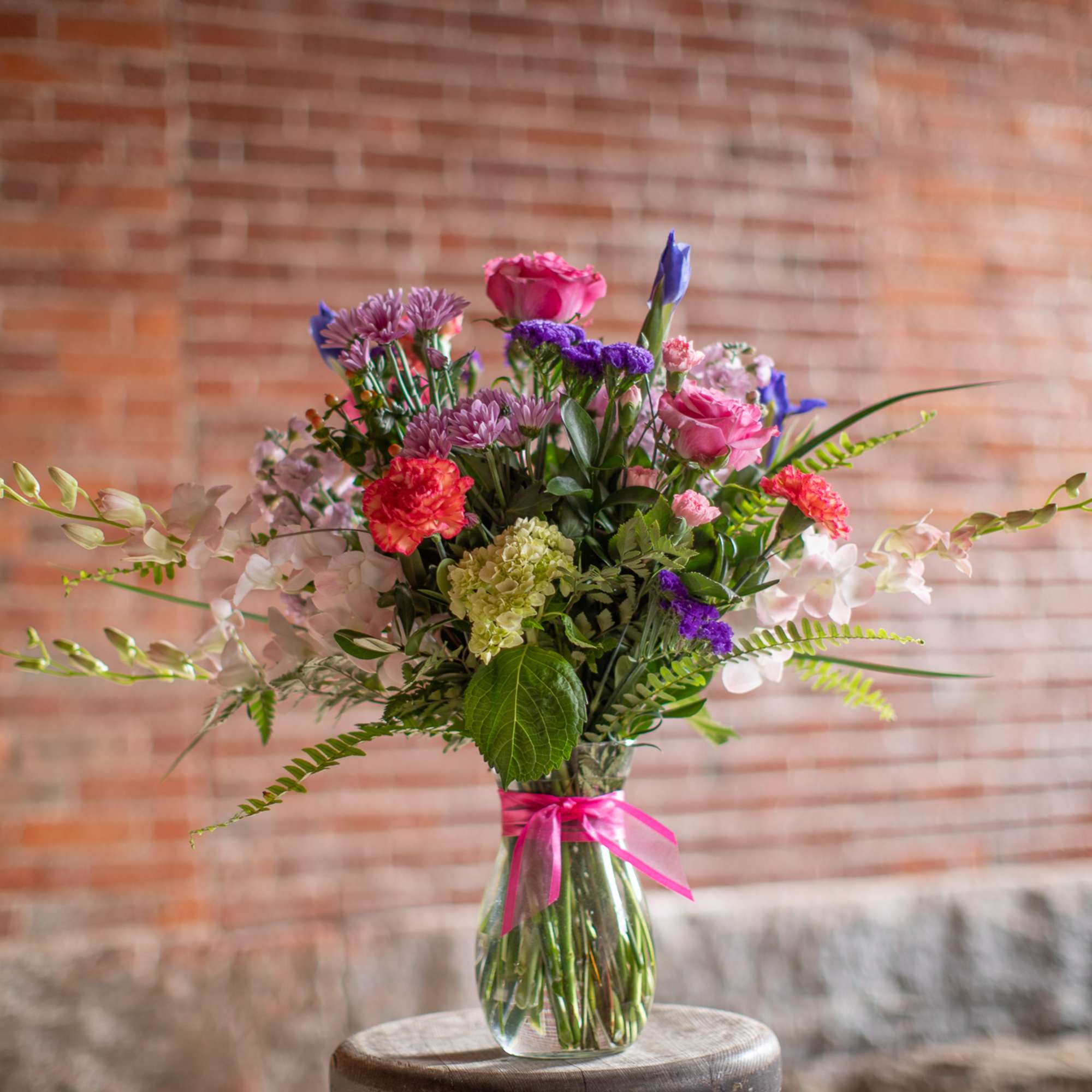 Mixed bouquet of pink, purple, and white flowers in a glass vase with a pink ribbon