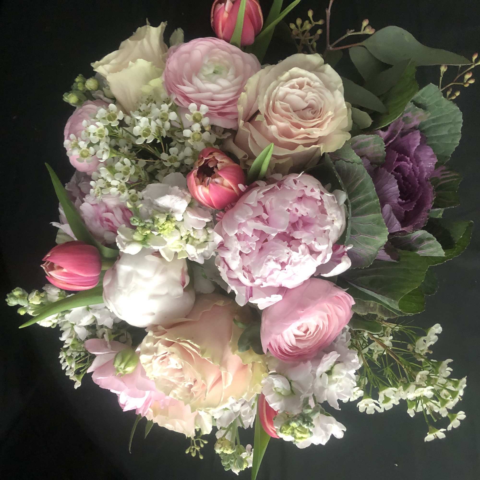 Hand-tied bouquet of pink peonies, roses, ranunculus, tulips, and white filler flowers on a dark background
