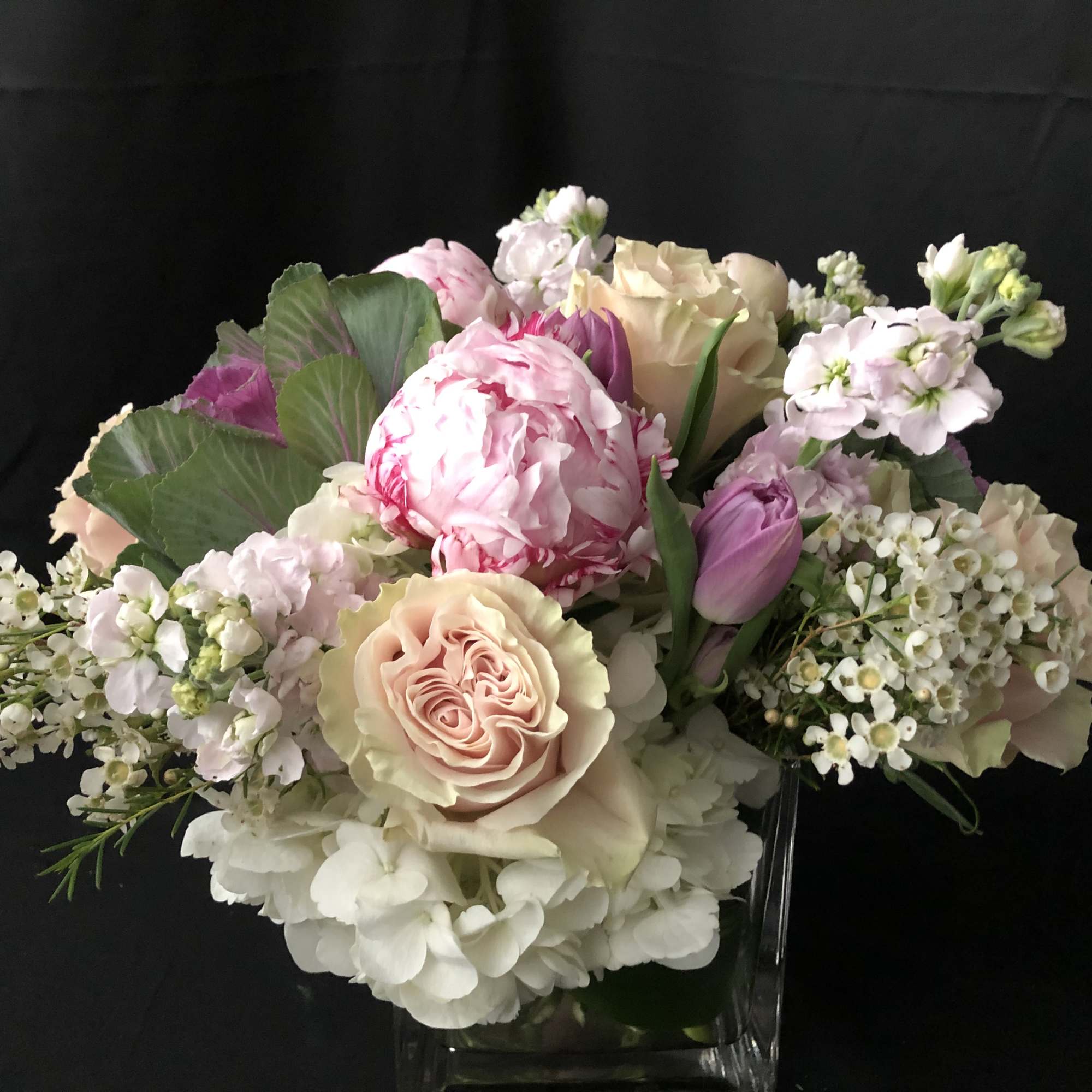 Low cube vase arrangement of pink peonies, peach roses, tulips, hydrangeas and white filler flowers.