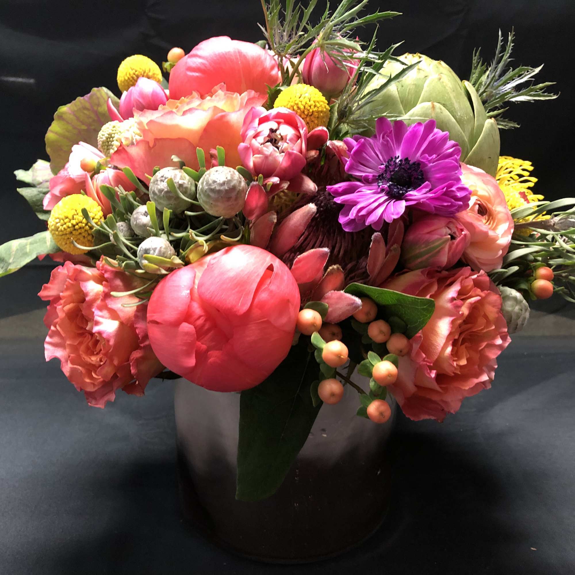Low arrangement of bright pink and orange blooms with yellow accents in a metallic cylinder vase