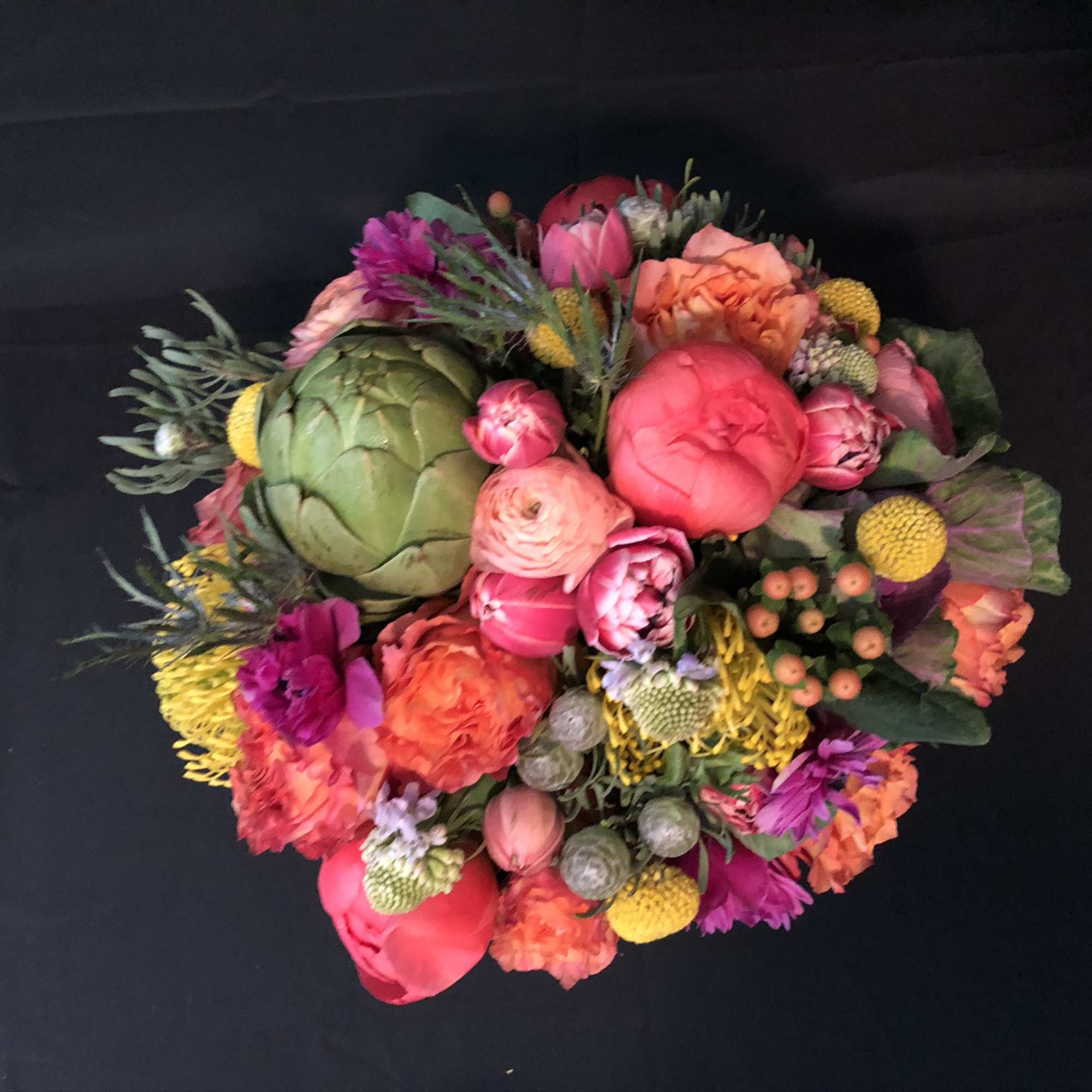 Round arrangement of bright pink, orange, and yellow flowers with a green artichoke accent on a black background
