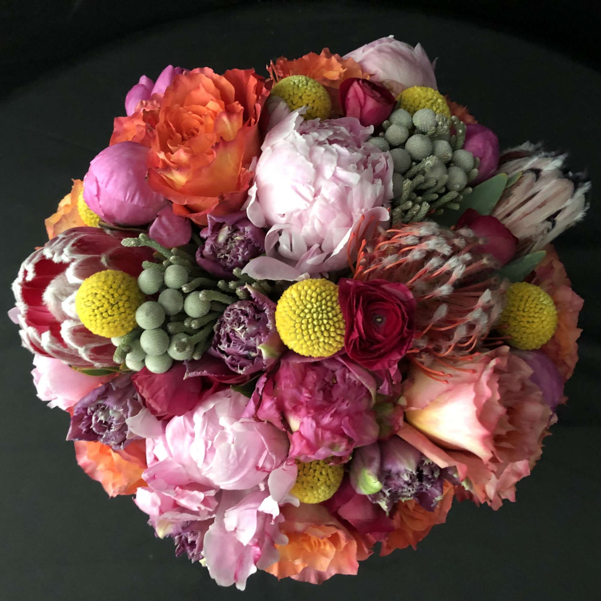 Round bouquet of pink peonies, roses, protea, and yellow ball flowers tightly clustered on black background