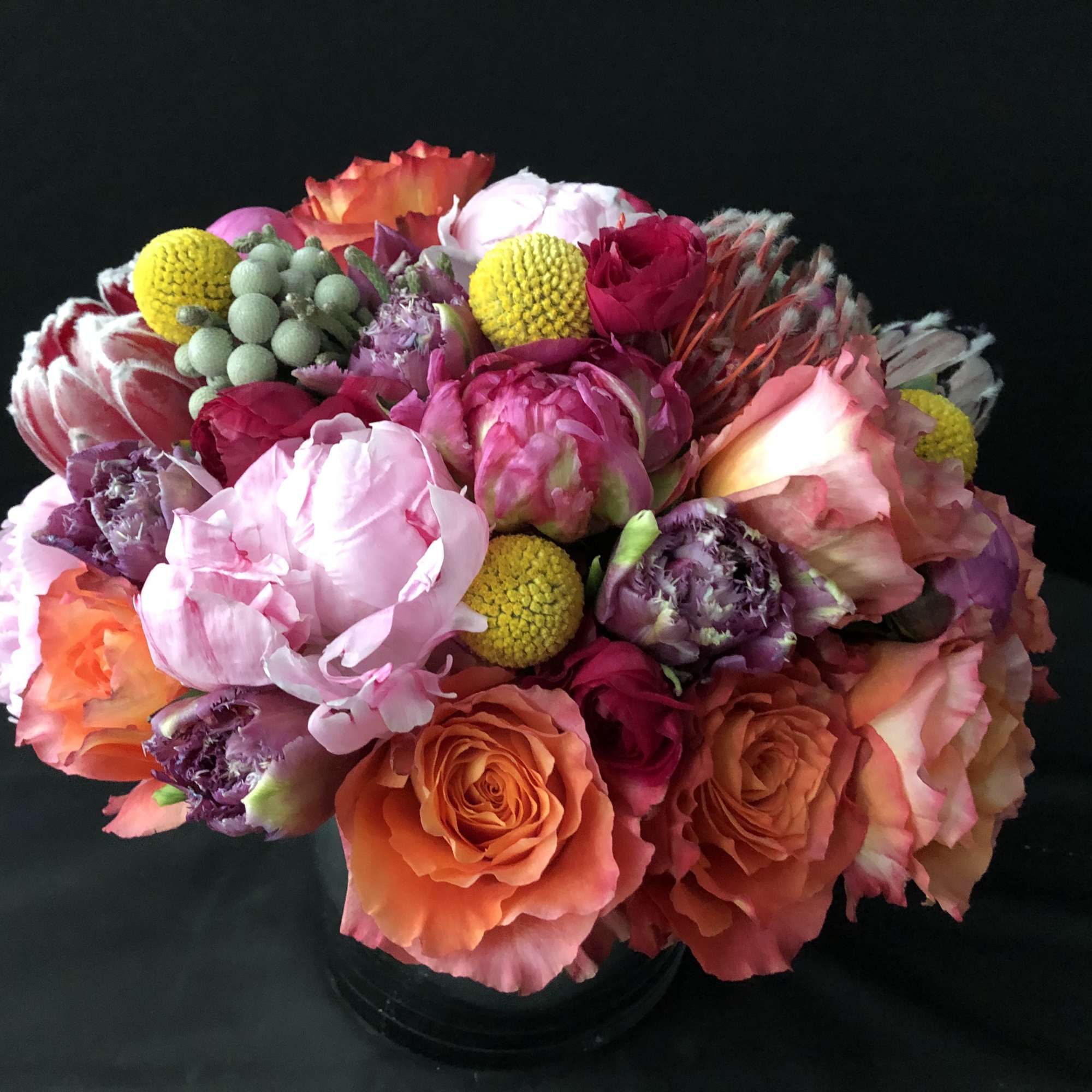 Round arrangement of pink peonies, orange roses, and yellow accent blooms in a black vase