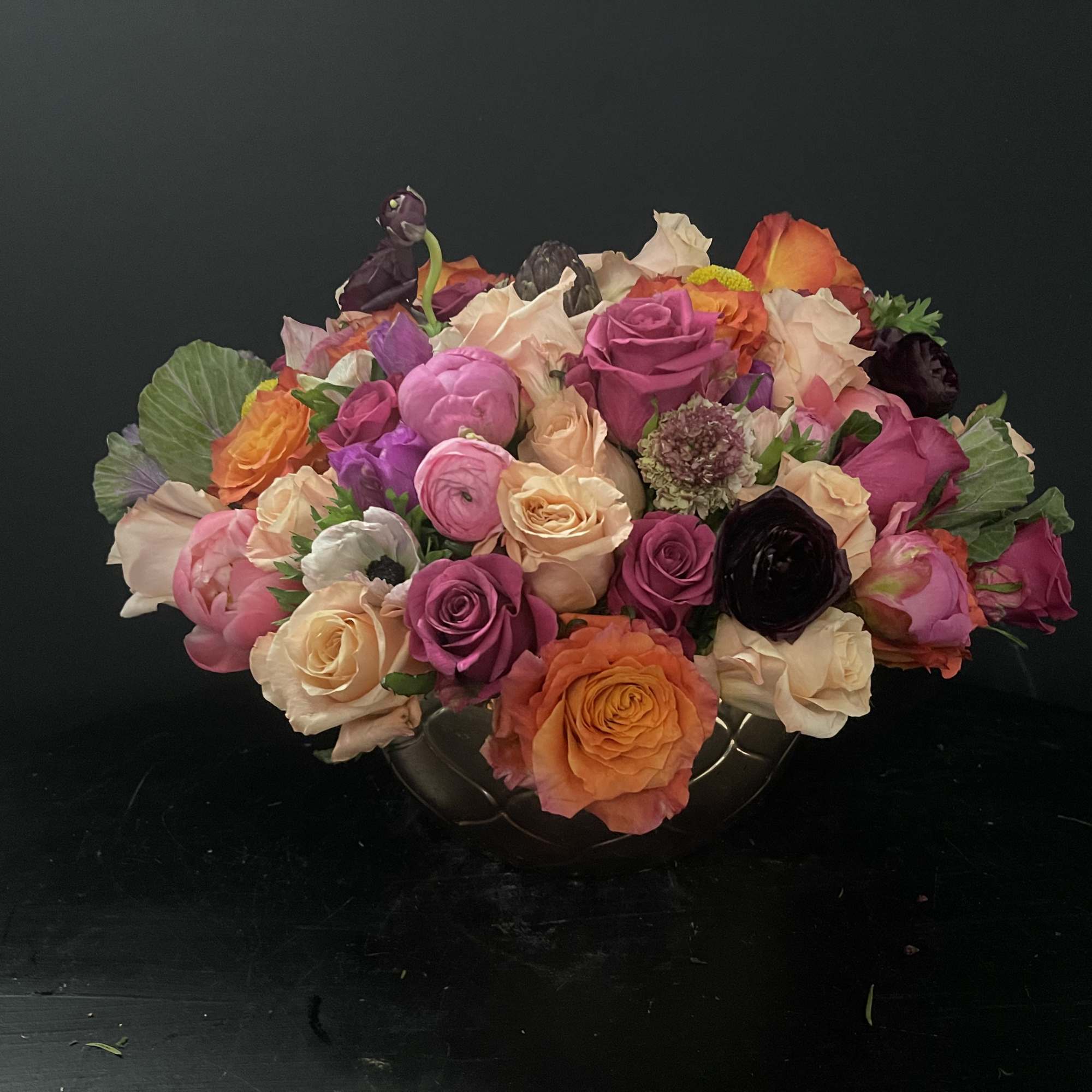 Low centerpiece of mixed pink, peach, orange, and purple roses and ranunculus in a round vase