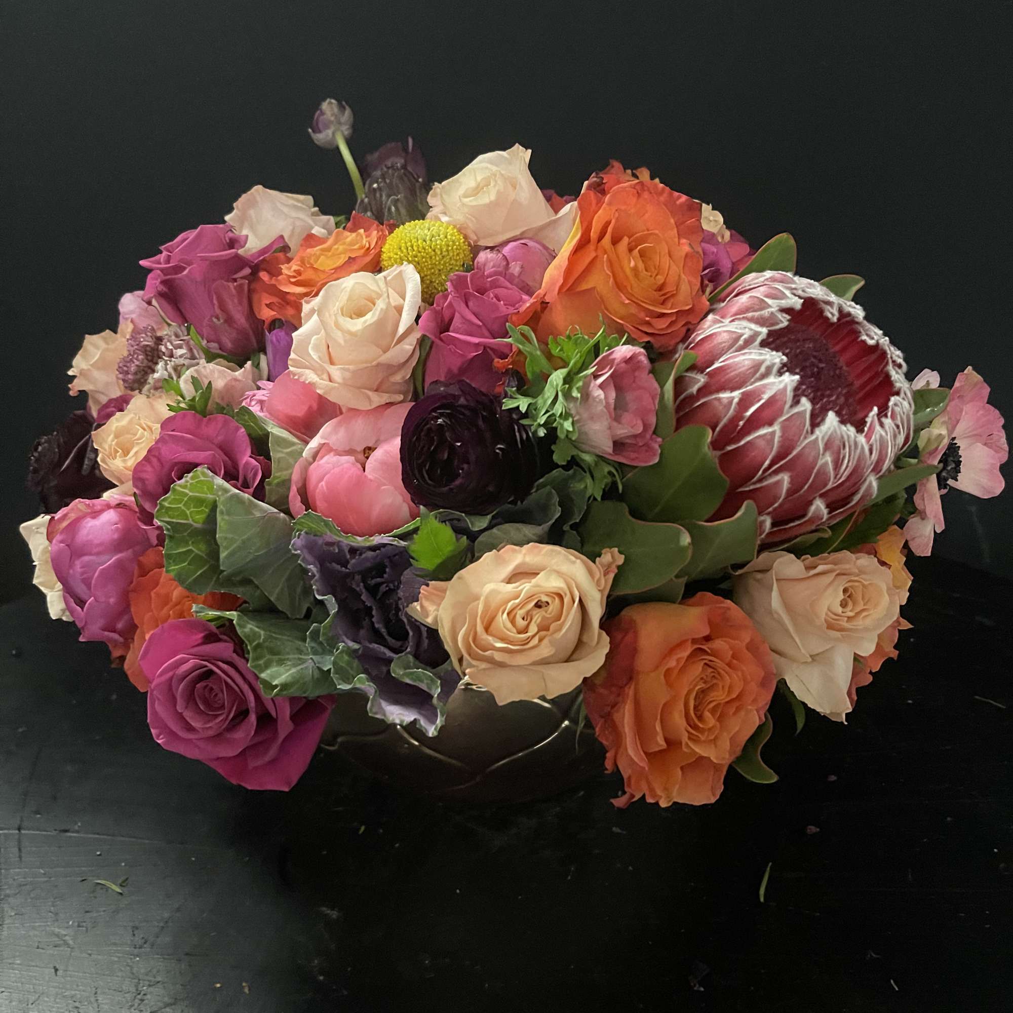 Low bowl arrangement of mixed roses, protea, and ranunculus in pink, peach, and orange tones.