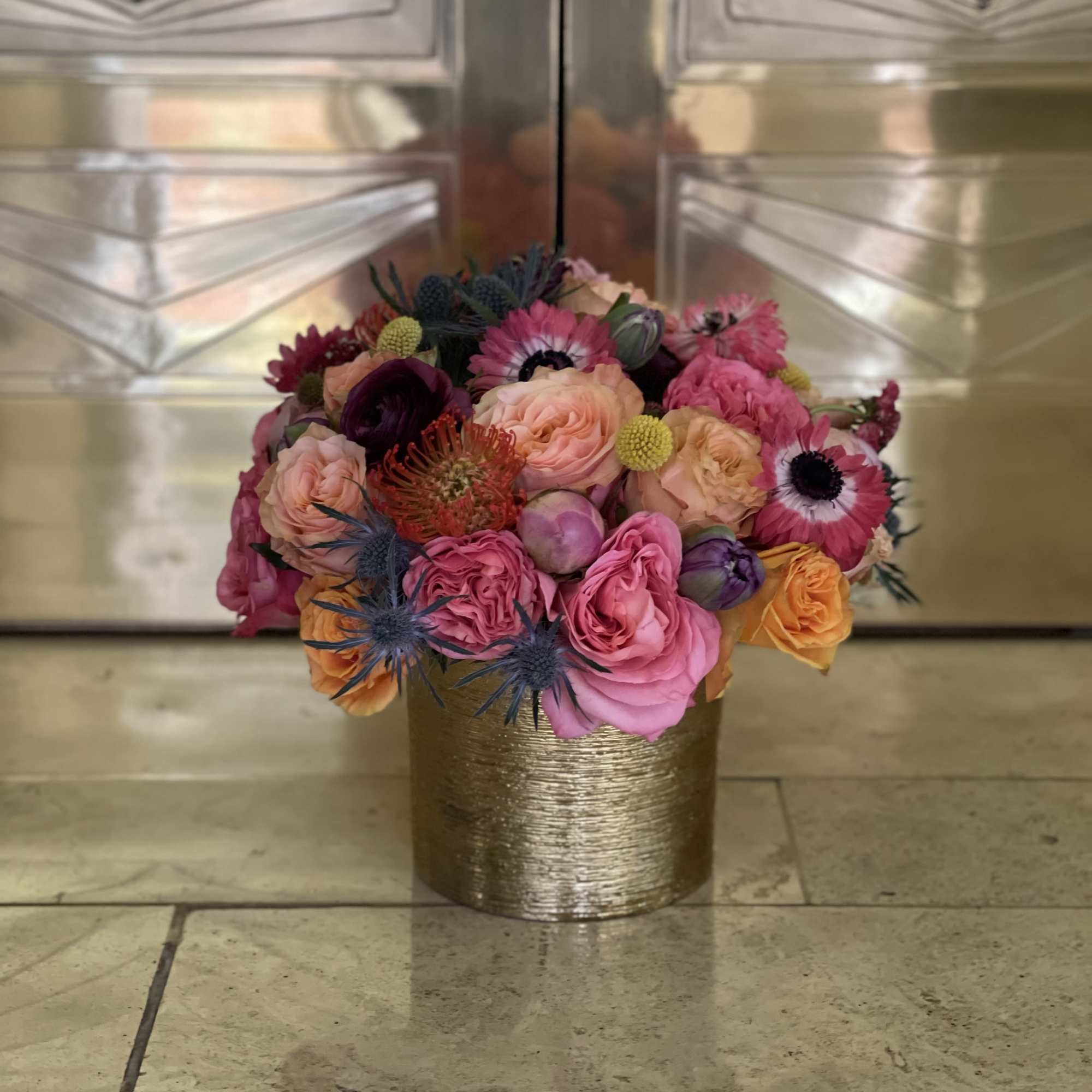 Bright pink and orange flower arrangement in a gold cylinder vase on a stone floor