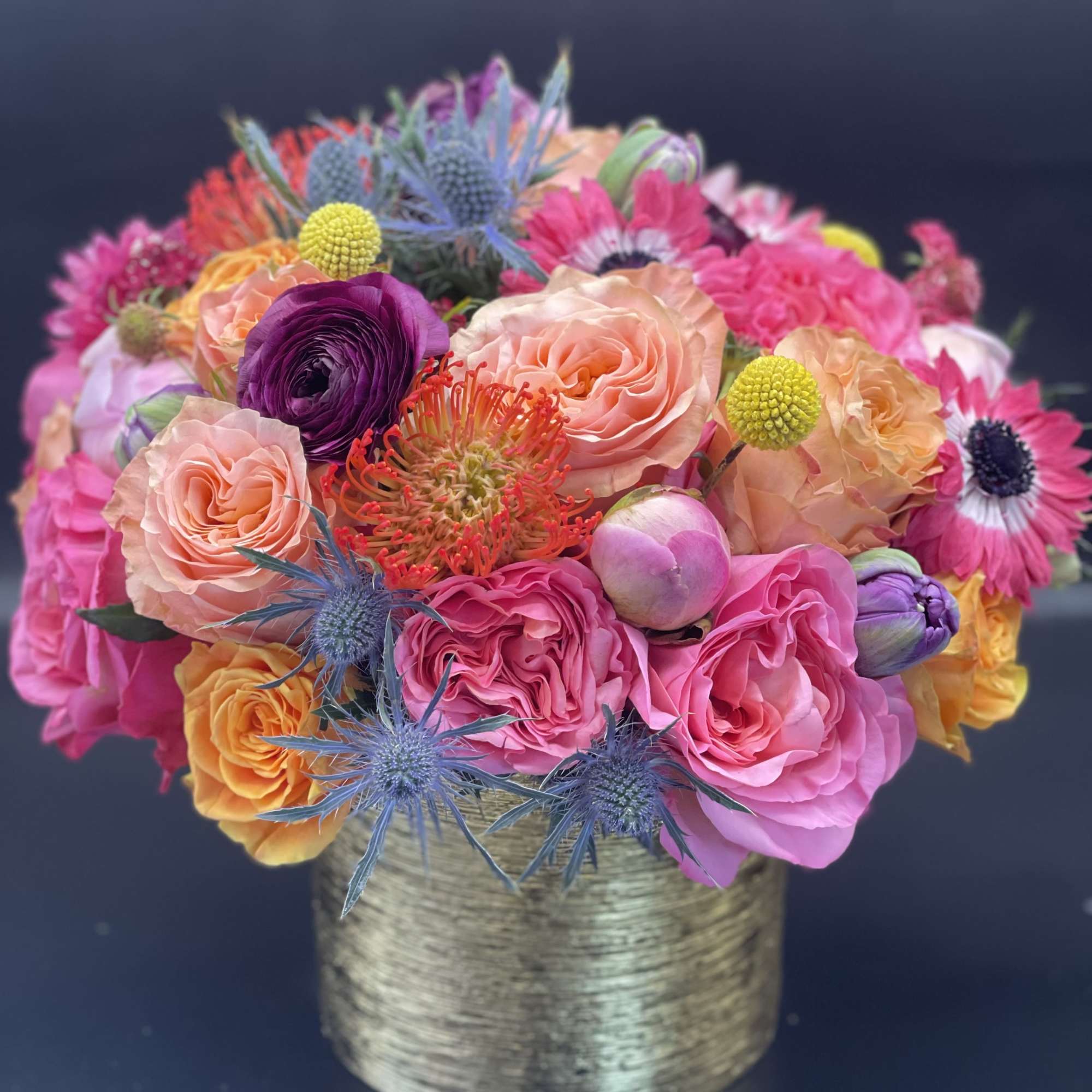 Low arrangement of bright pink, peach, and orange flowers with blue accents in a gold cylinder vase