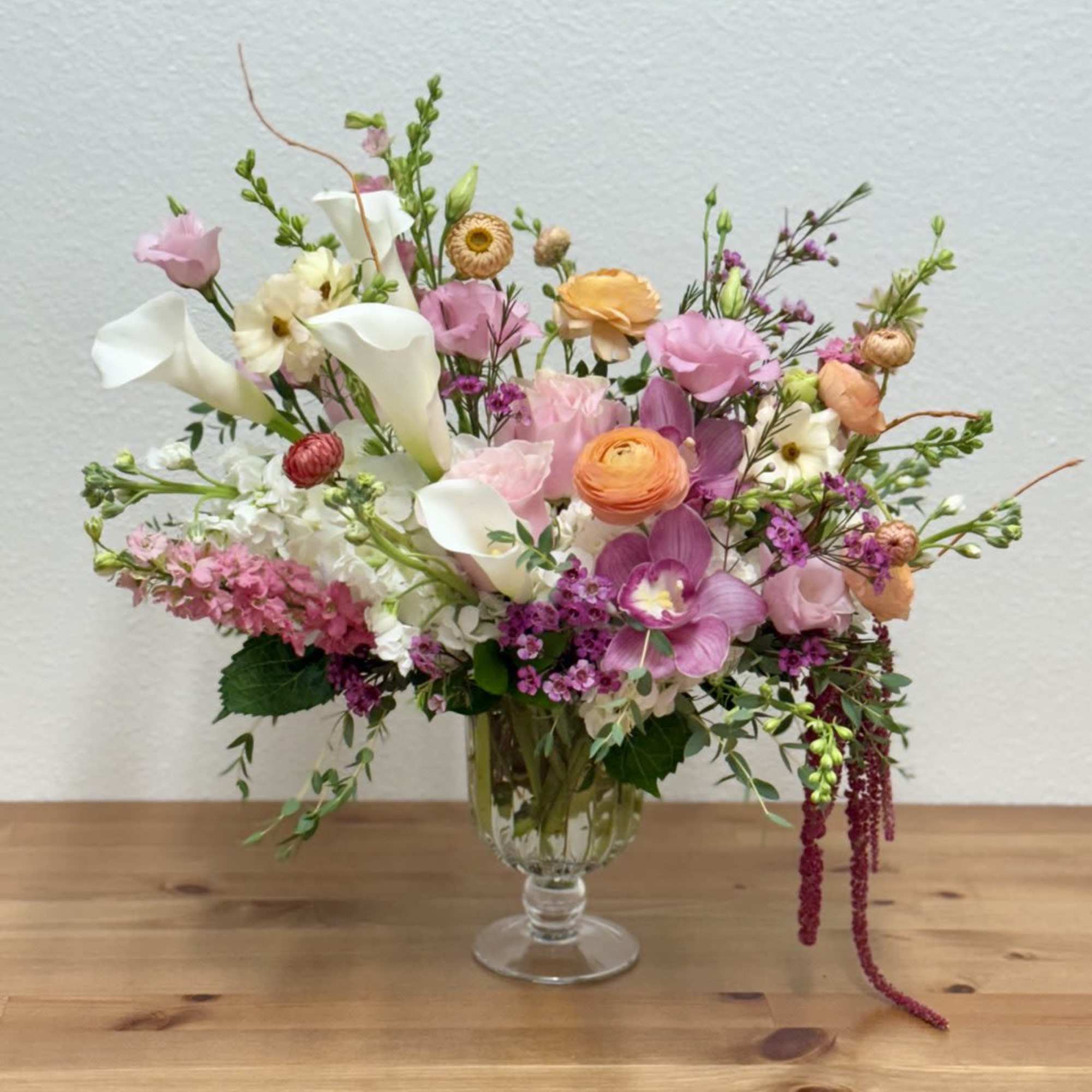 Asymmetrical pastel arrangement with calla lilies, ranunculus, orchids, and mixed blooms in a clear glass vase