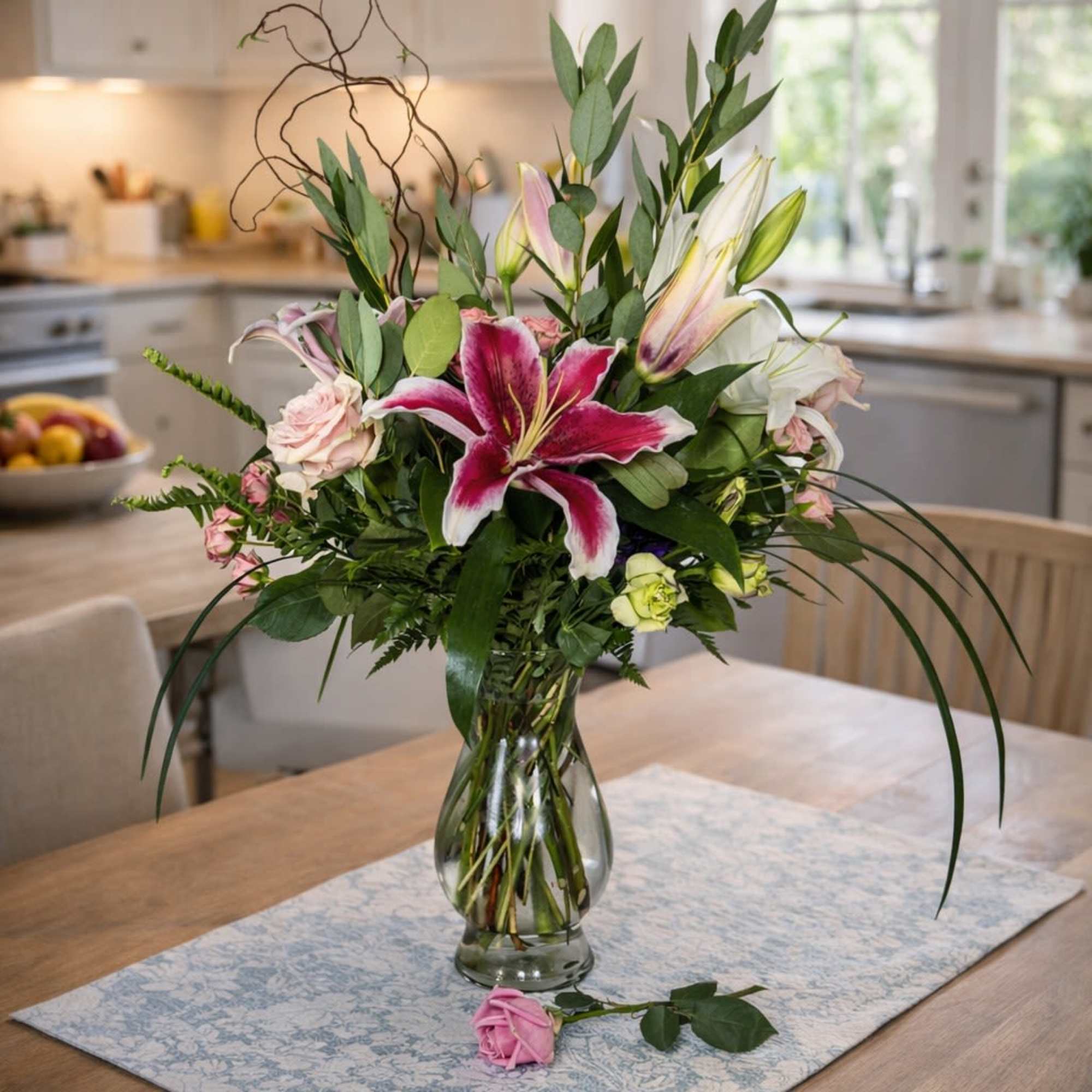 Tall arrangement of pink lilies and pale roses in a clear glass vase on a wooden table