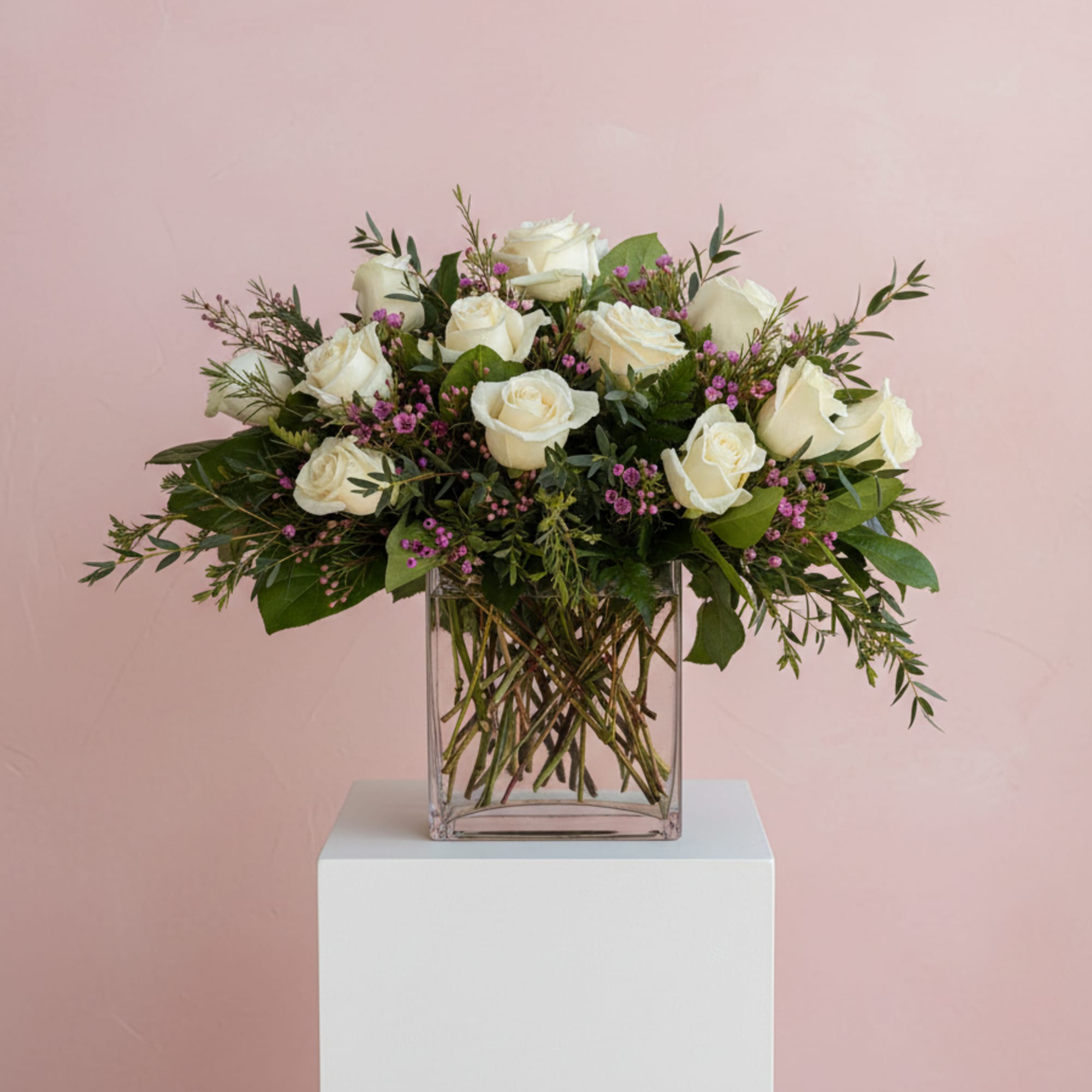 Arrangement of white roses with pink filler flowers in a clear glass cube vase against a pink background
