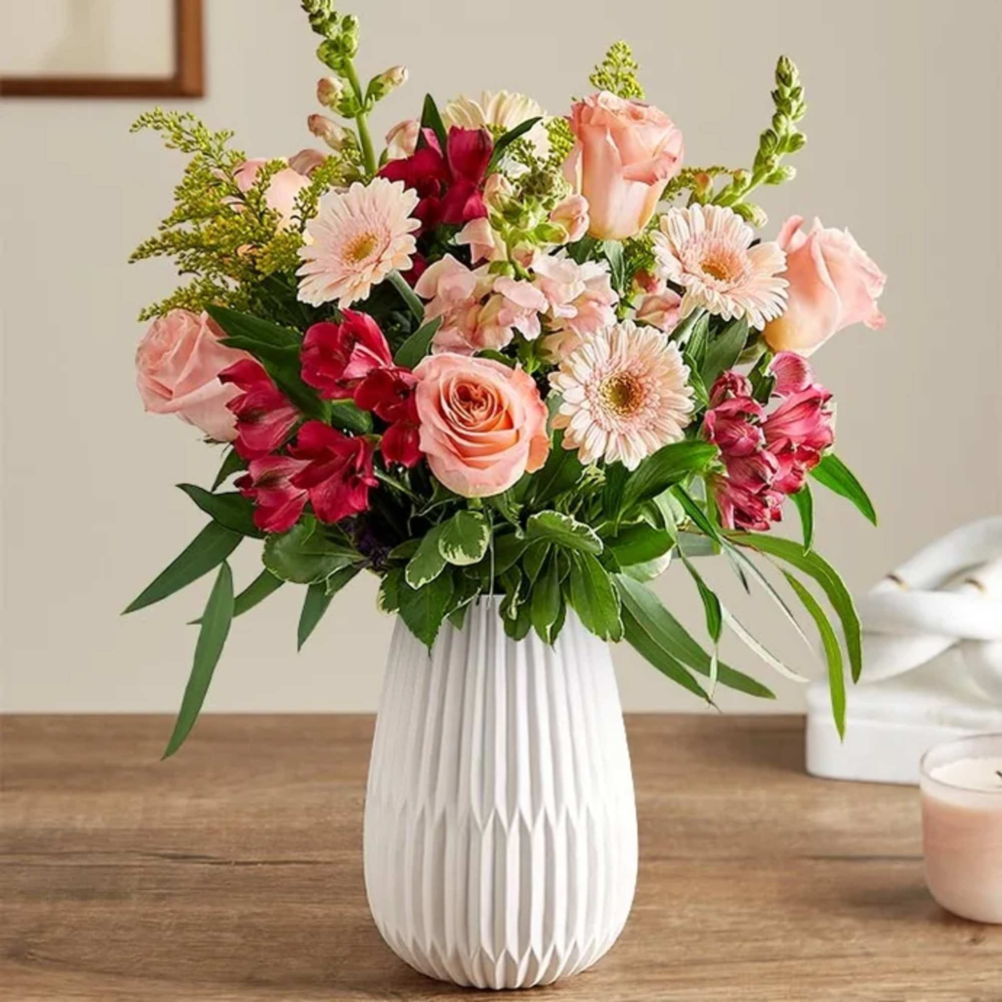 Mixed bouquet of pink roses, gerbera daisies, and red blooms in a white vase.