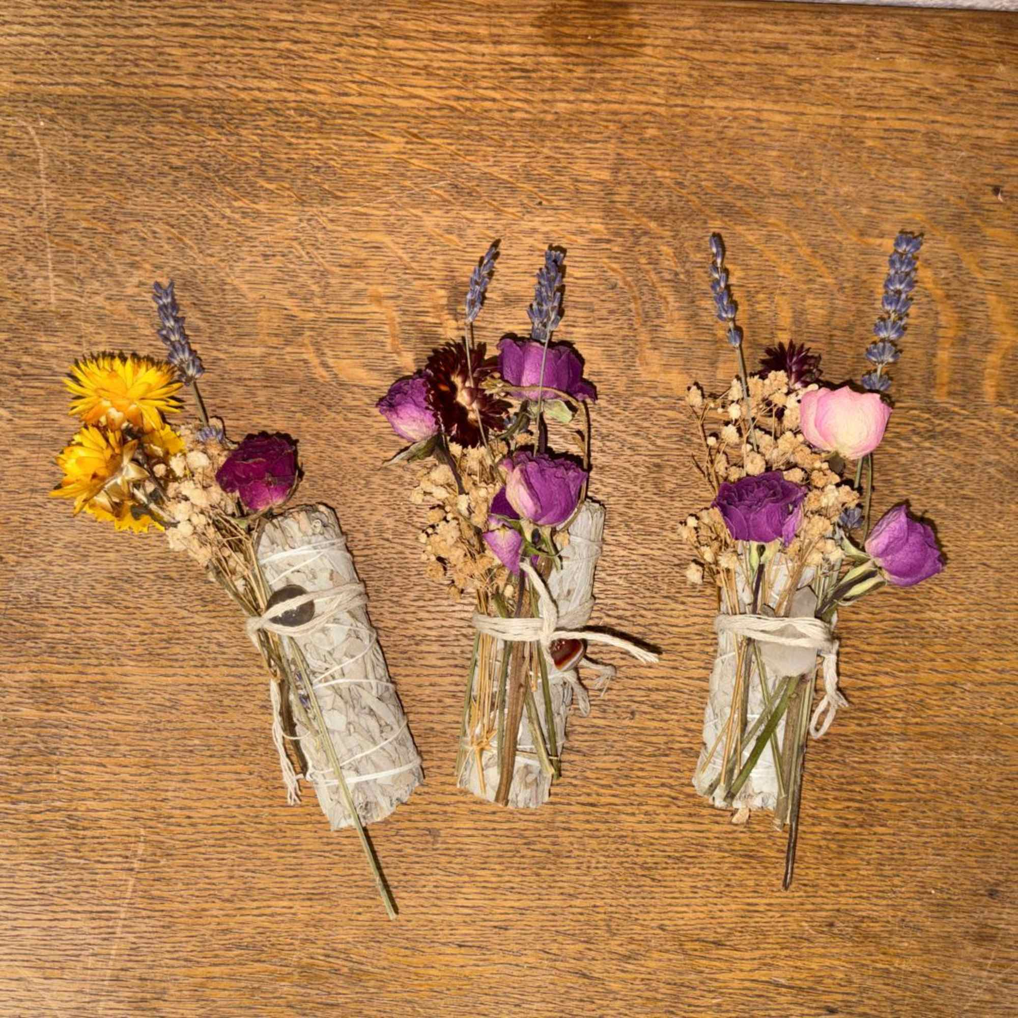 Three small dried flower and sage bundles tied with twine on a wooden surface