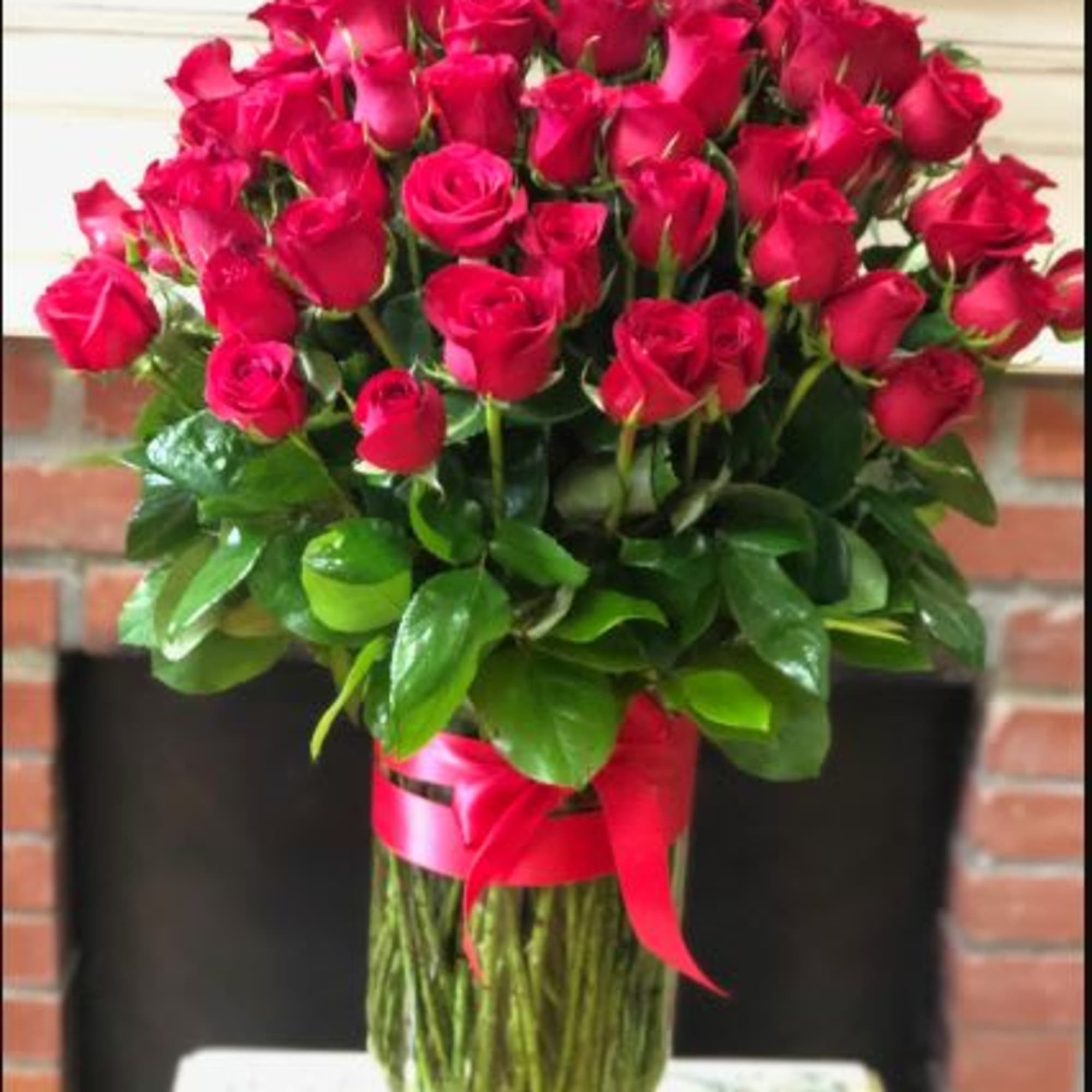 Large arrangement of many red roses in a tall clear glass vase with a red ribbon bow.