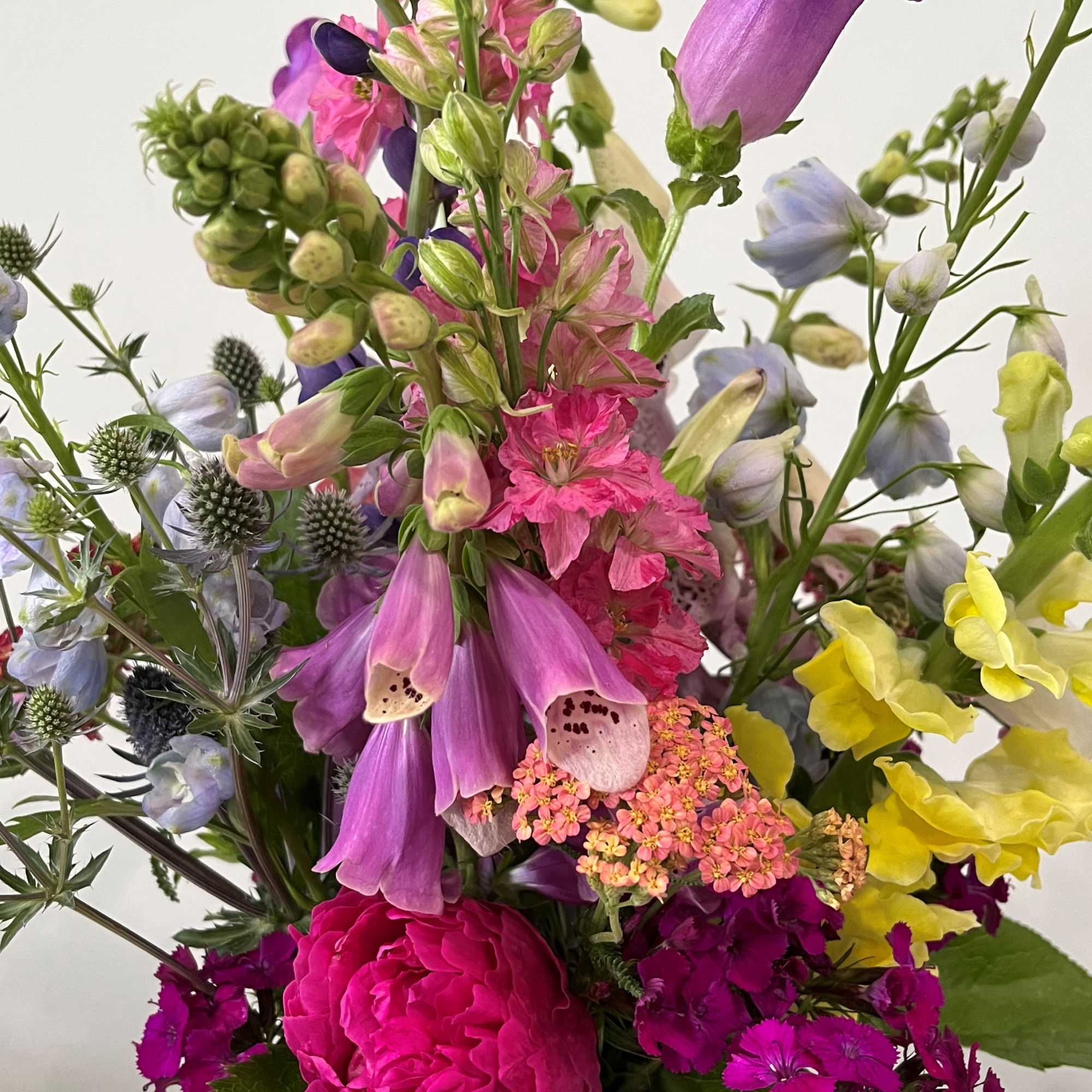 Tall mixed bouquet of pink peony, purple bells, yellow snapdragons, and other garden flowers in a glass vase