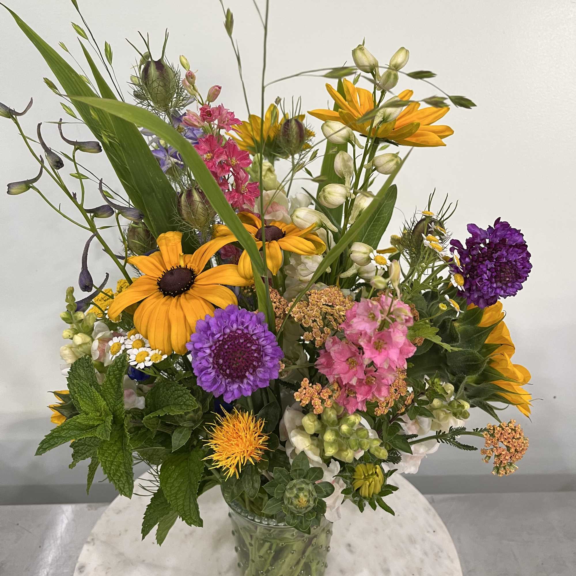 Tall mixed wildflower arrangement in a green glass vase on a round marble table