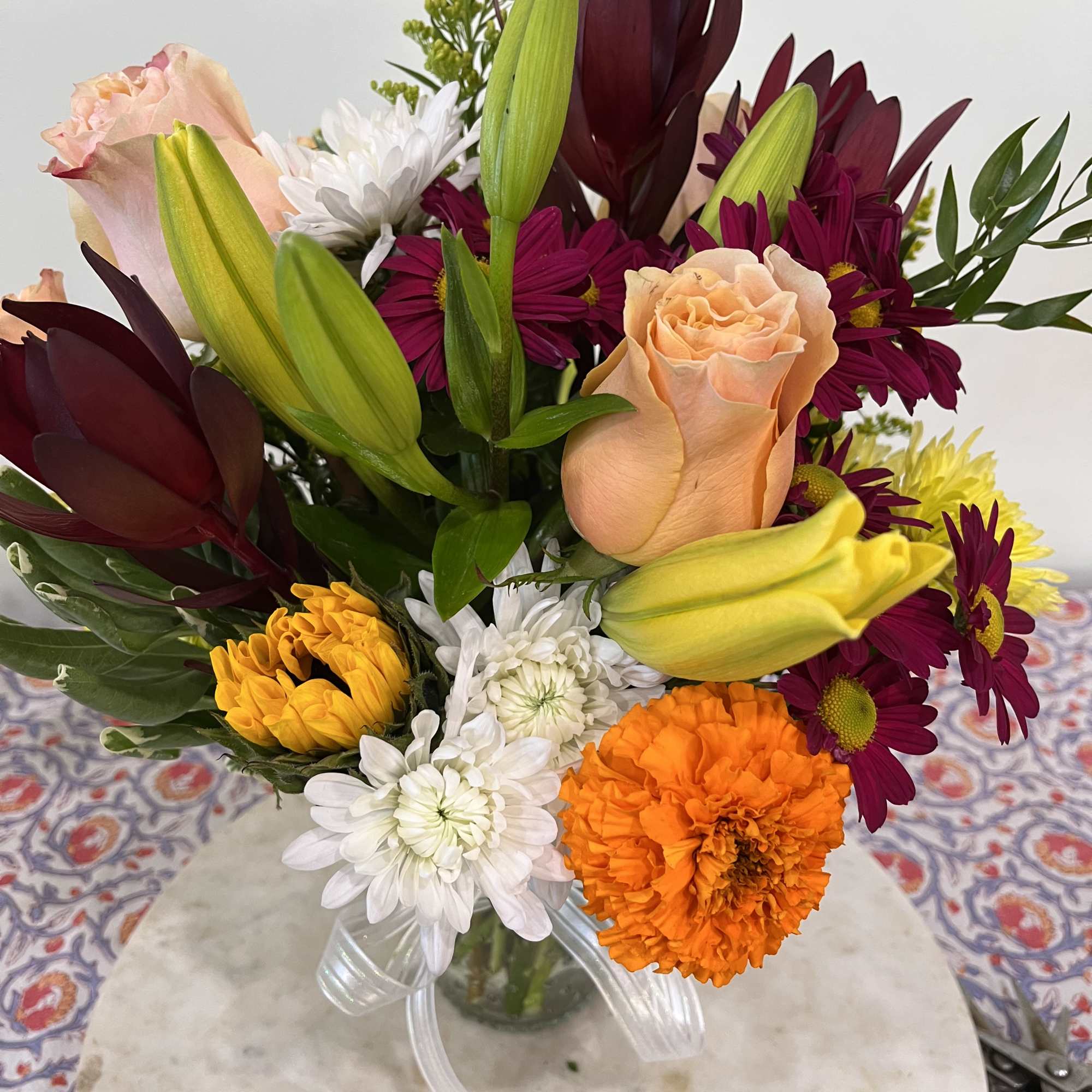 Mixed bouquet of peach roses, yellow lilies, marigolds, and white and purple mums in a clear glass vase
