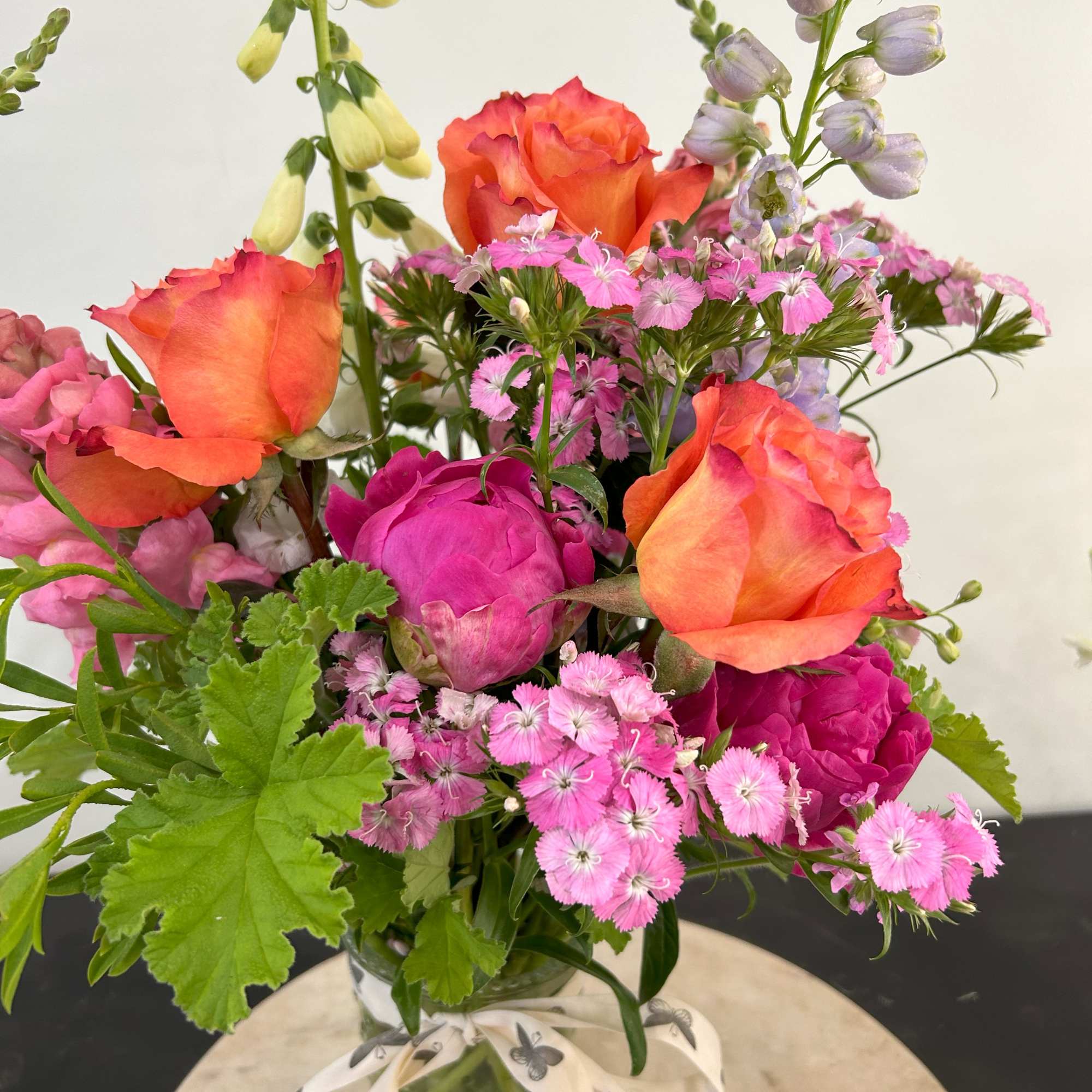 Bright bouquet of orange roses, hot pink blooms, and pale accents in a glass jar with ribbon.