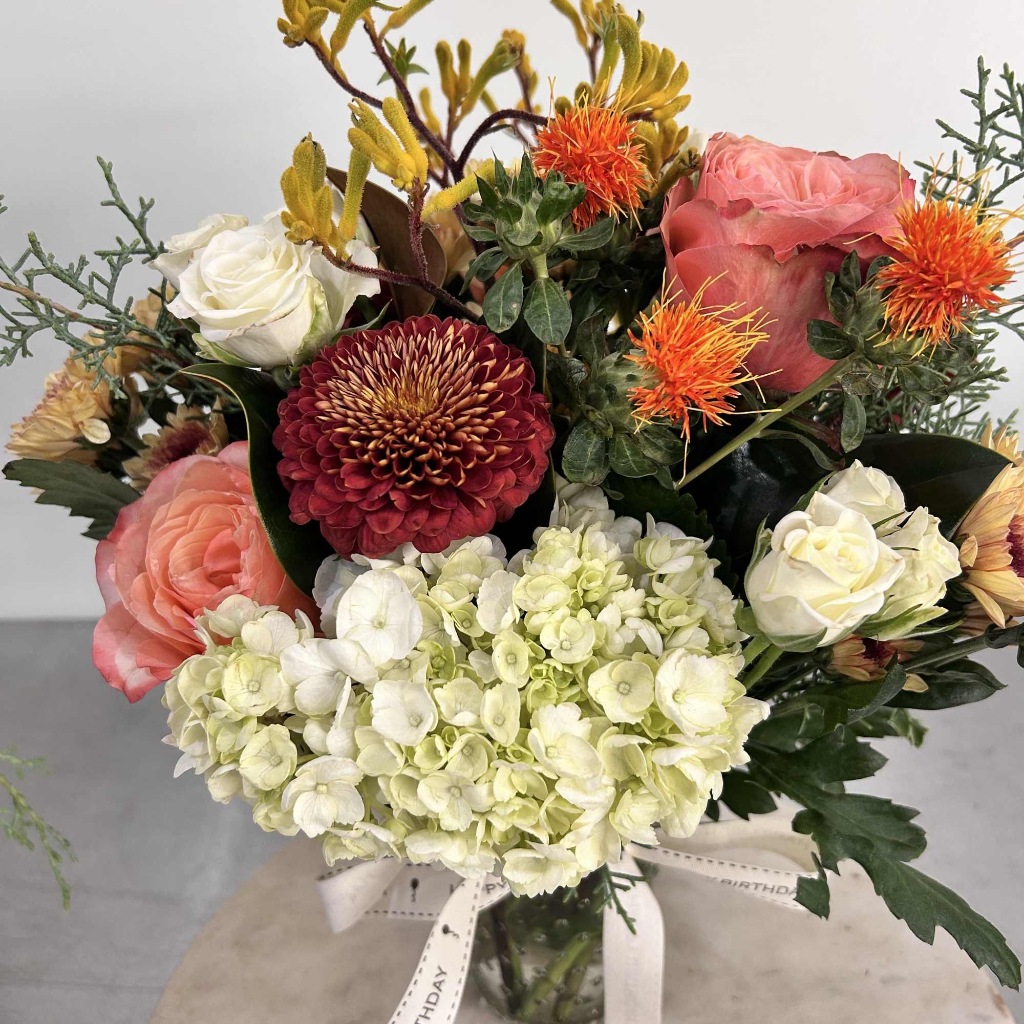 Mixed arrangement of peach roses, white hydrangeas, and red mum in a glass vase with Happy Birthday ribbon.