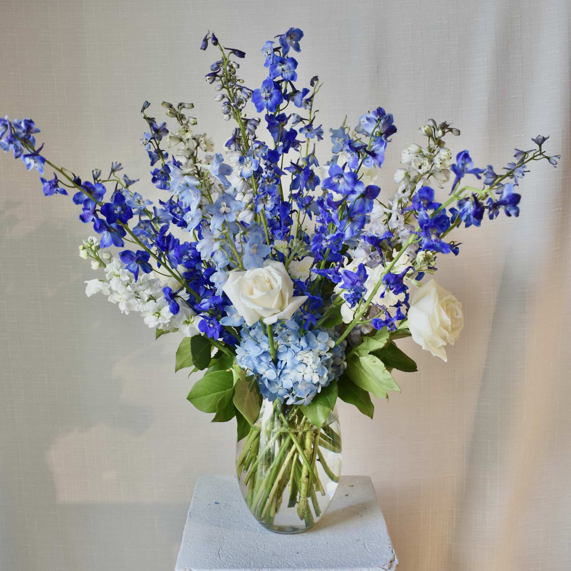 Tall blue and white flower arrangement with roses, hydrangea, and delphinium in a clear glass vase