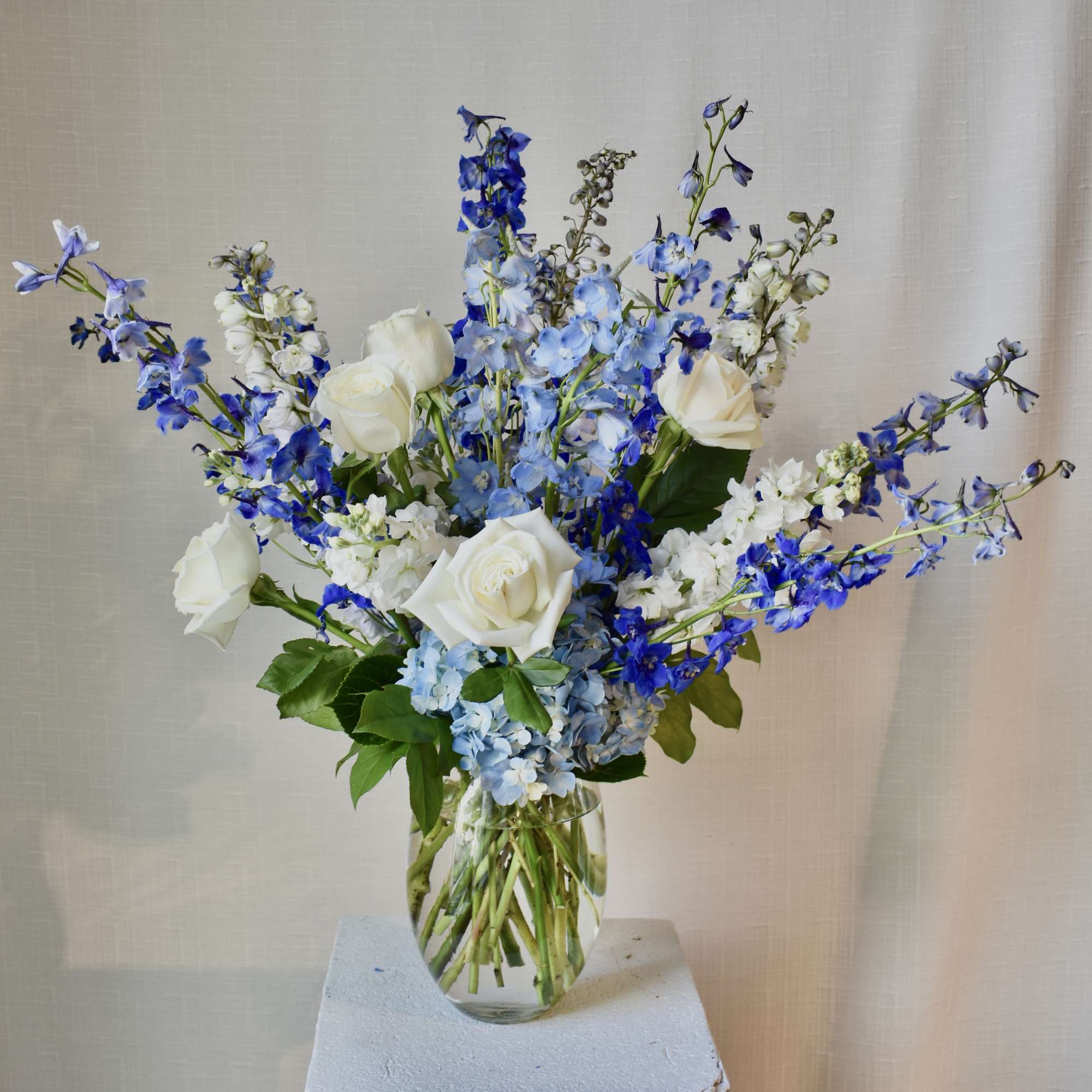 Tall arrangement of white roses and blue delphinium in a clear glass vase.