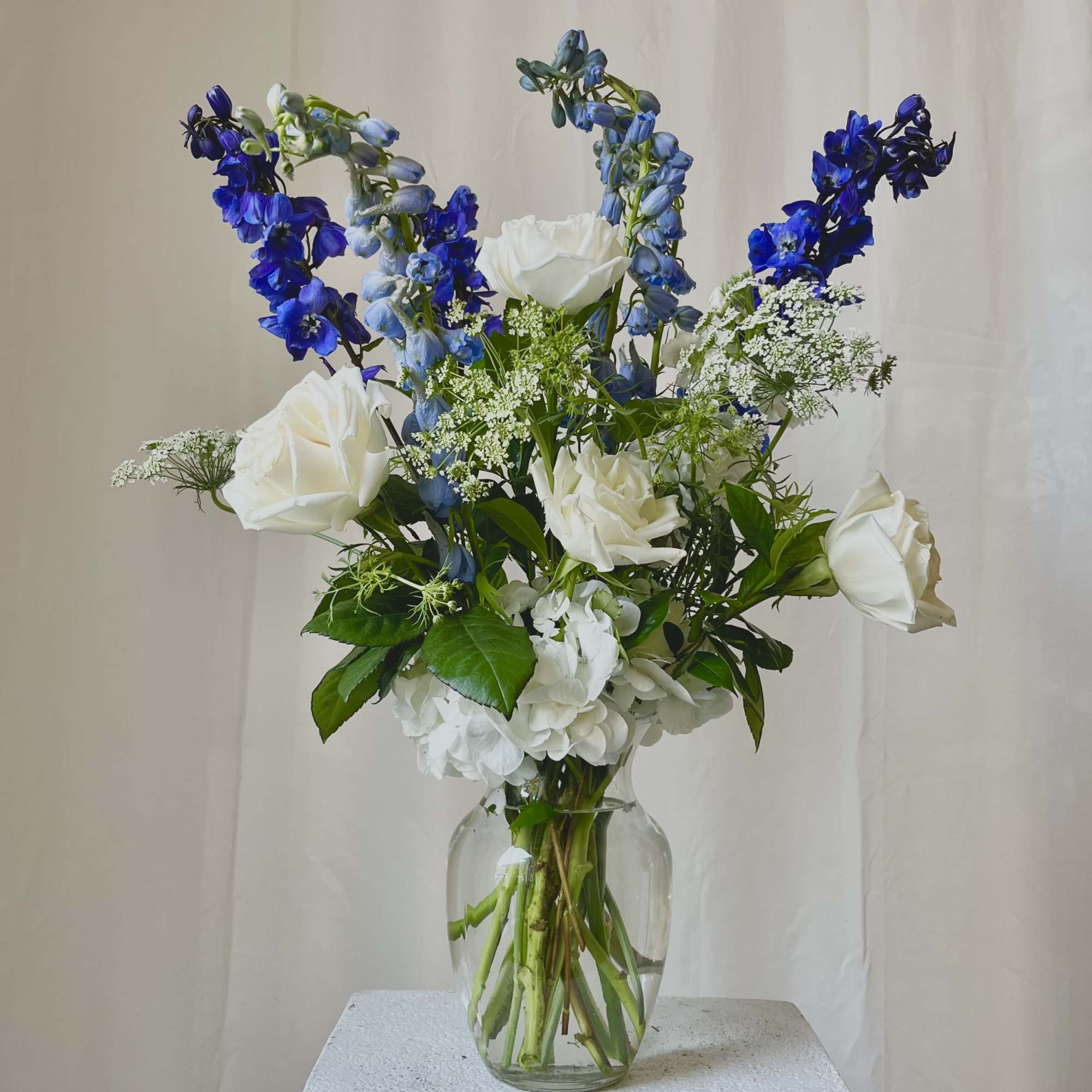 Tall arrangement of white roses and hydrangea with blue delphinium in a clear glass vase